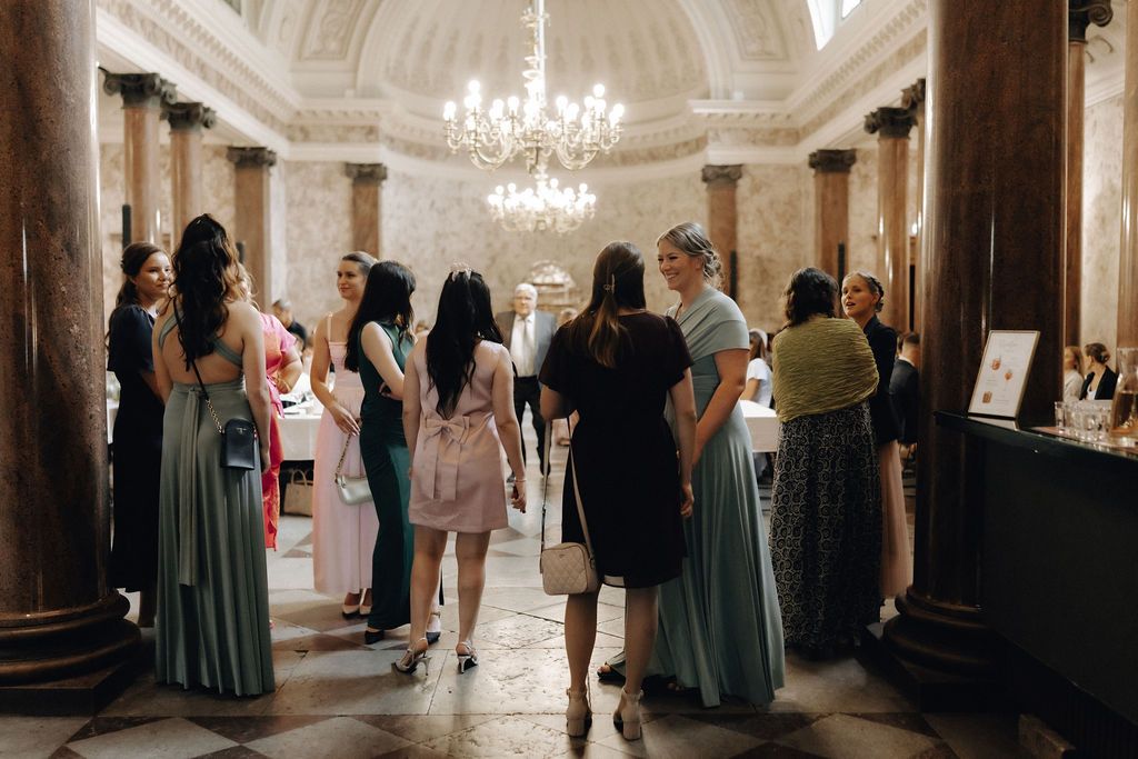 Wedding guests in sage green gowns mingle in a grand marble-columned reception hall.