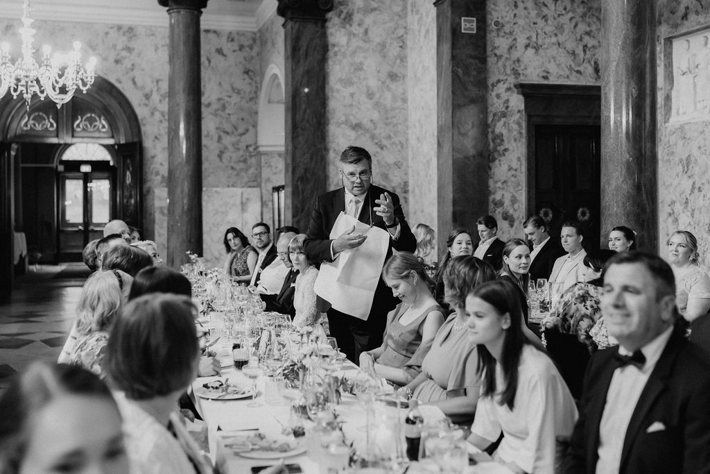 Man giving speech at elegant wedding reception dinner in ornate marble hall with chandelier.