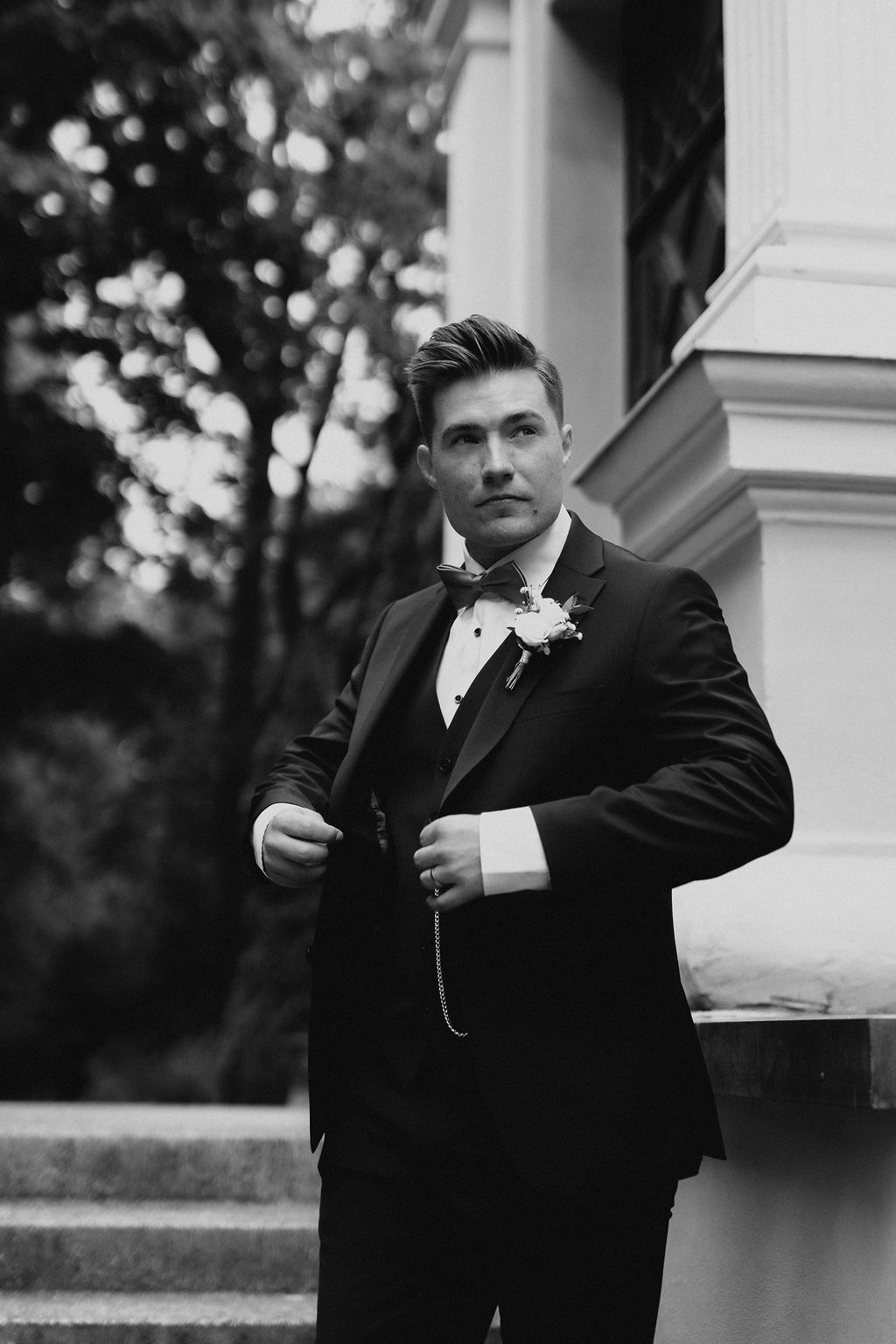 Groom in black tuxedo and bow tie adjusting jacket near stone pillar, black and white portrait.