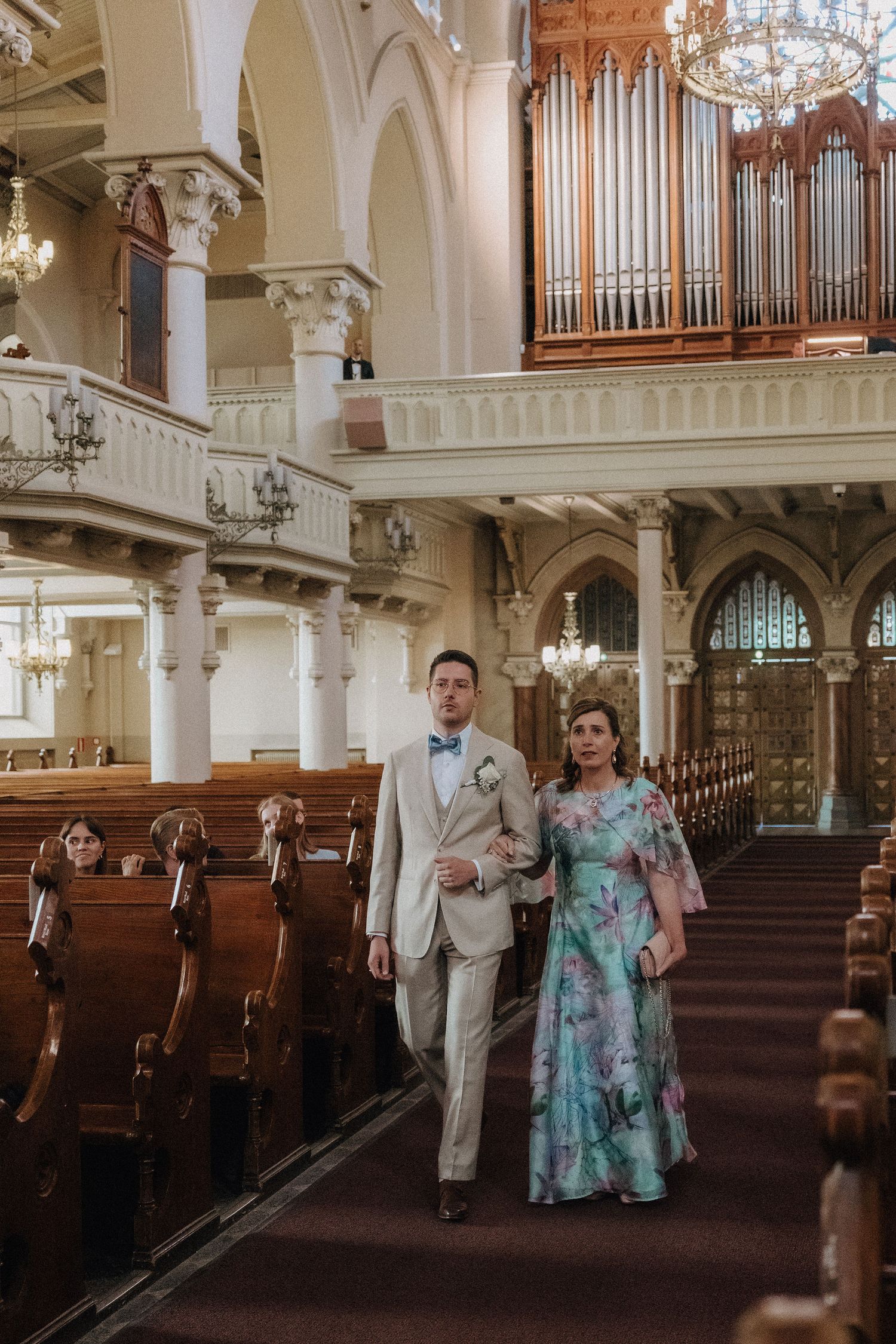 Groom in beige suit escorts woman in floral gown down ornate church aisle during wedding.