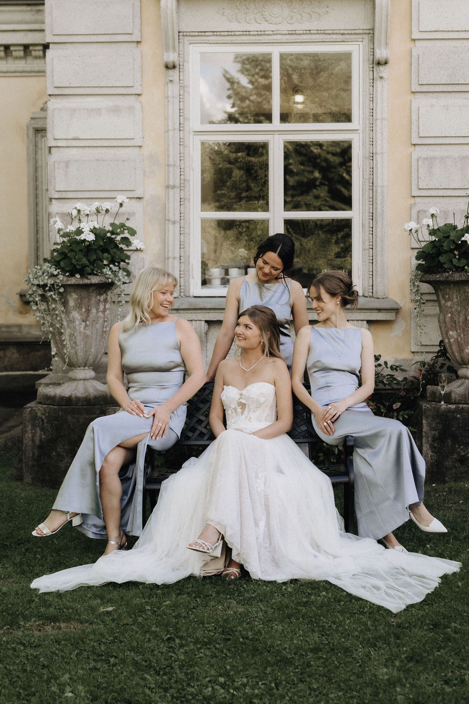 Bride in strapless lace gown seated on bench surrounded by three bridesmaids in blue-grey satin dresses outdoors.