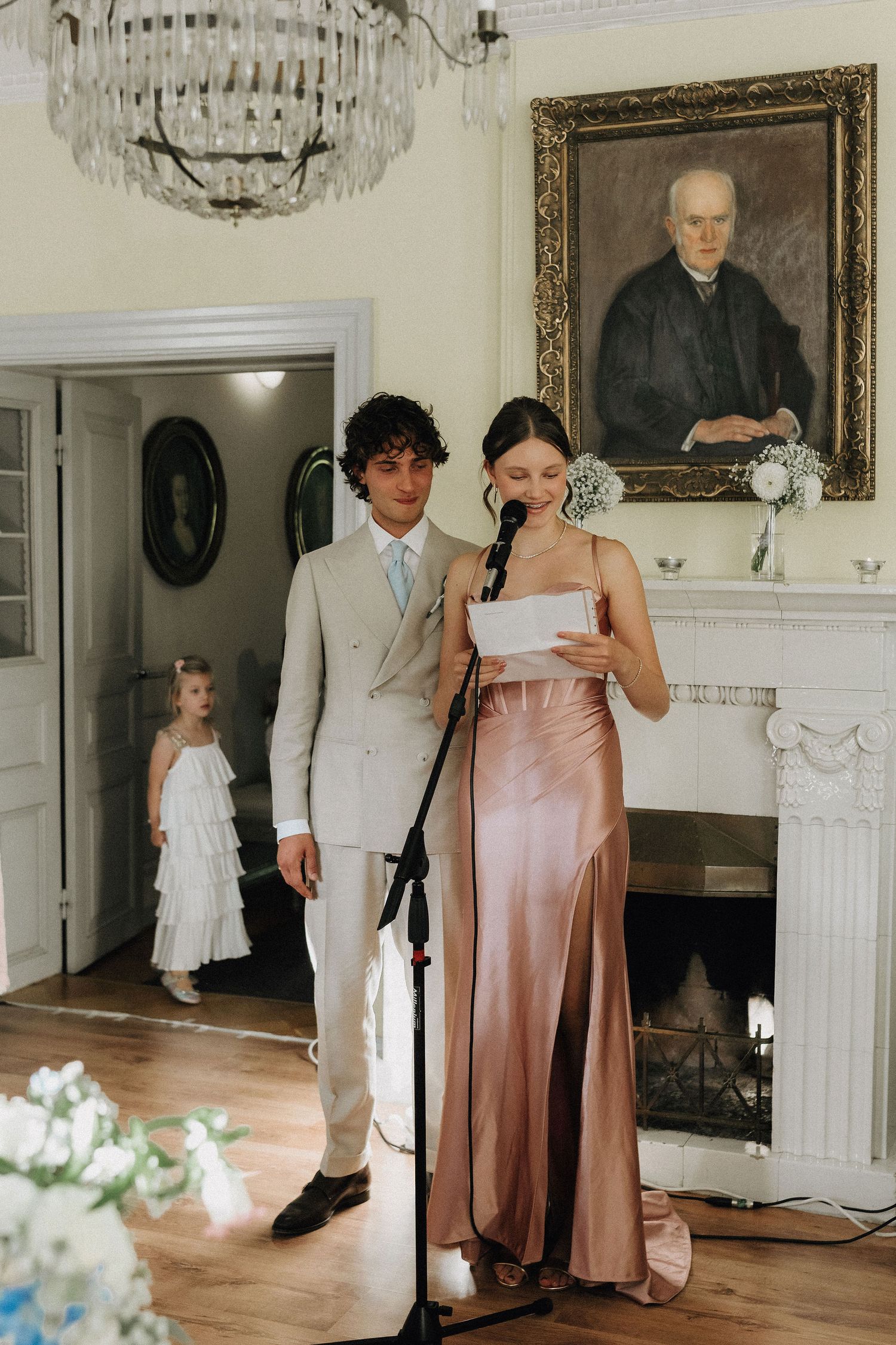 Bridesmaid in rose gold satin gown reading wedding speech at microphone in elegant manor room.