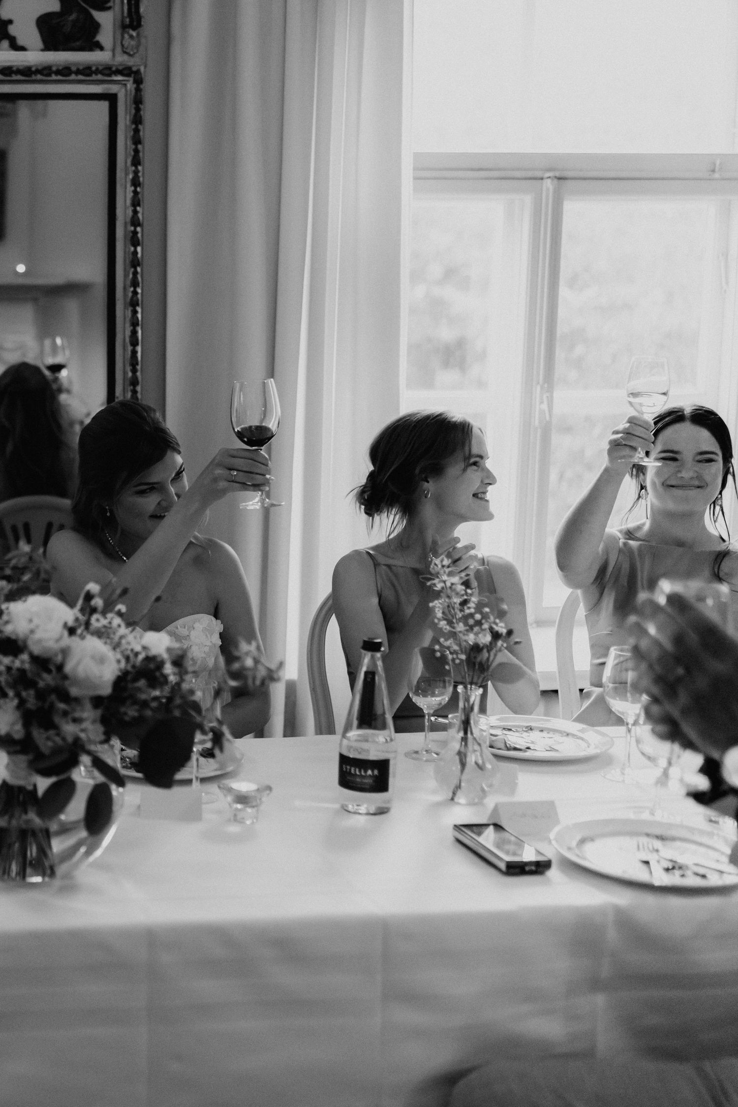 Three bridesmaids raising glasses in a toast at wedding reception table, black and white photo.