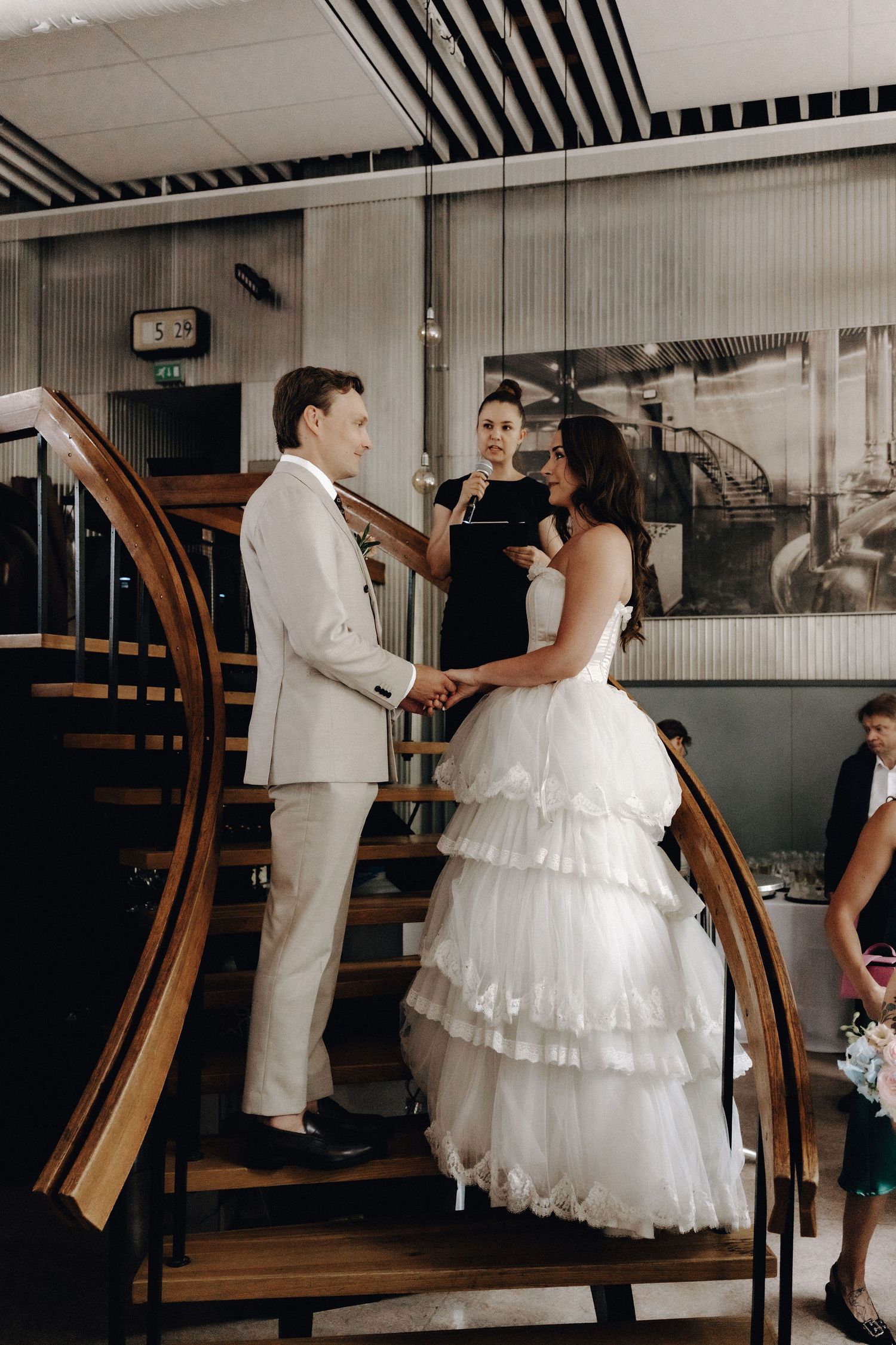 Bride and groom exchanging vows indoors near a wooden spiral staircase with an officiant present.
