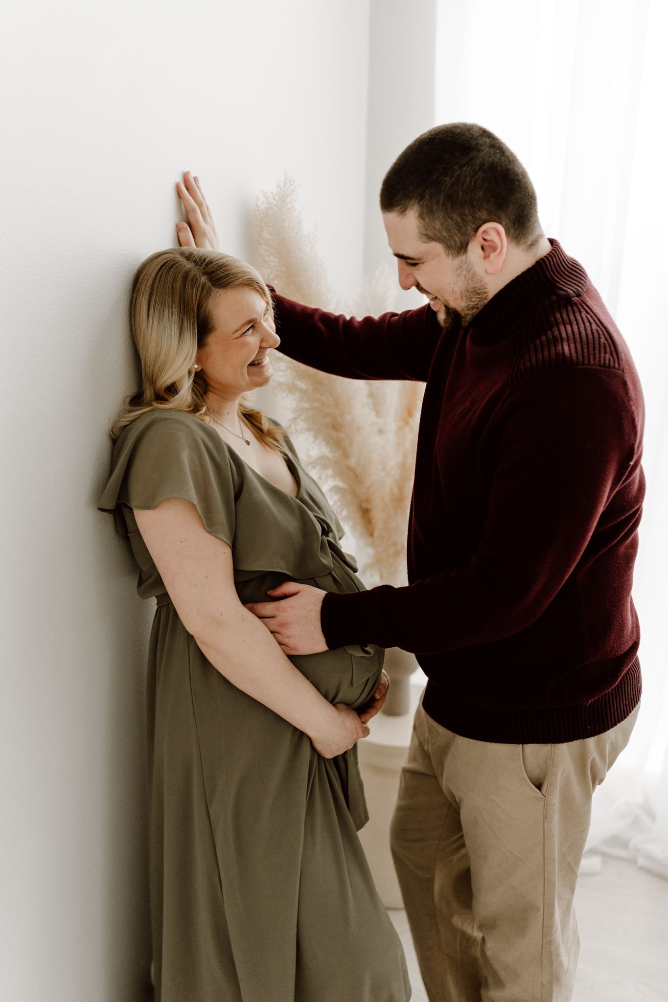 Smiling pregnant couple leaning against wall, man in burgundy sweater holding womans face, woman in olive maternity dress.