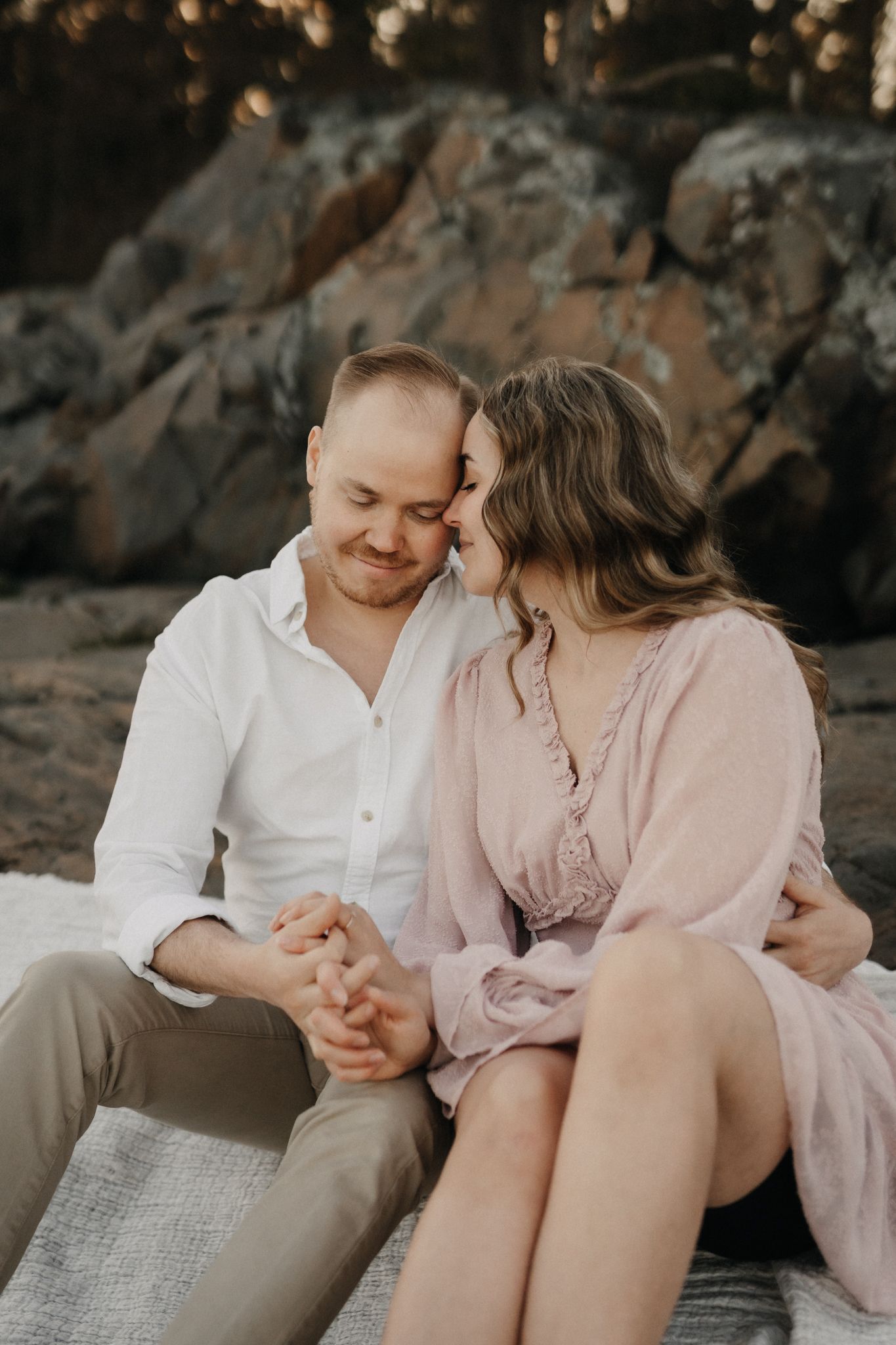 Couple sitting close on white blanket by rocky shore, holding hands and leaning together tenderly.