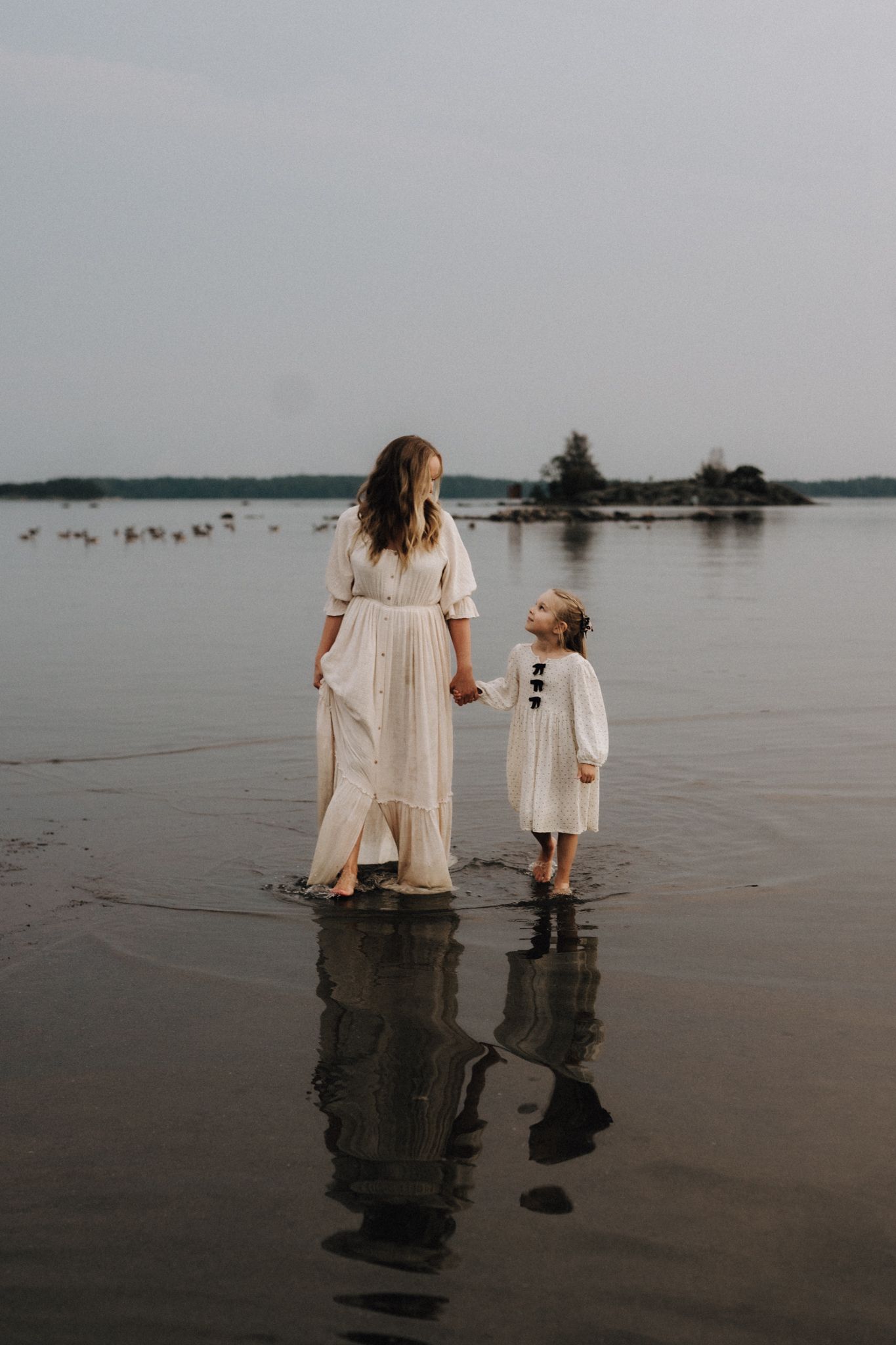Mother and young daughter in matching white dresses standing in shallow lake water, looking at each other.