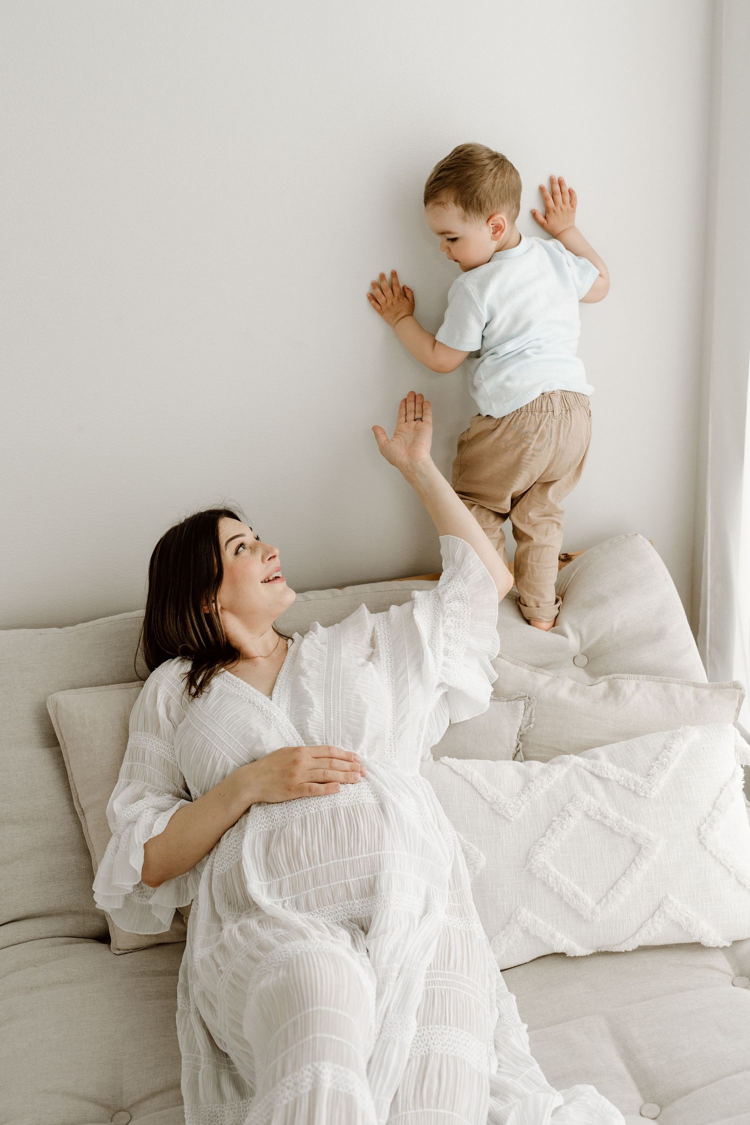 A pregnant woman in white sits on a bed smiling as her toddler son stands against the wall.