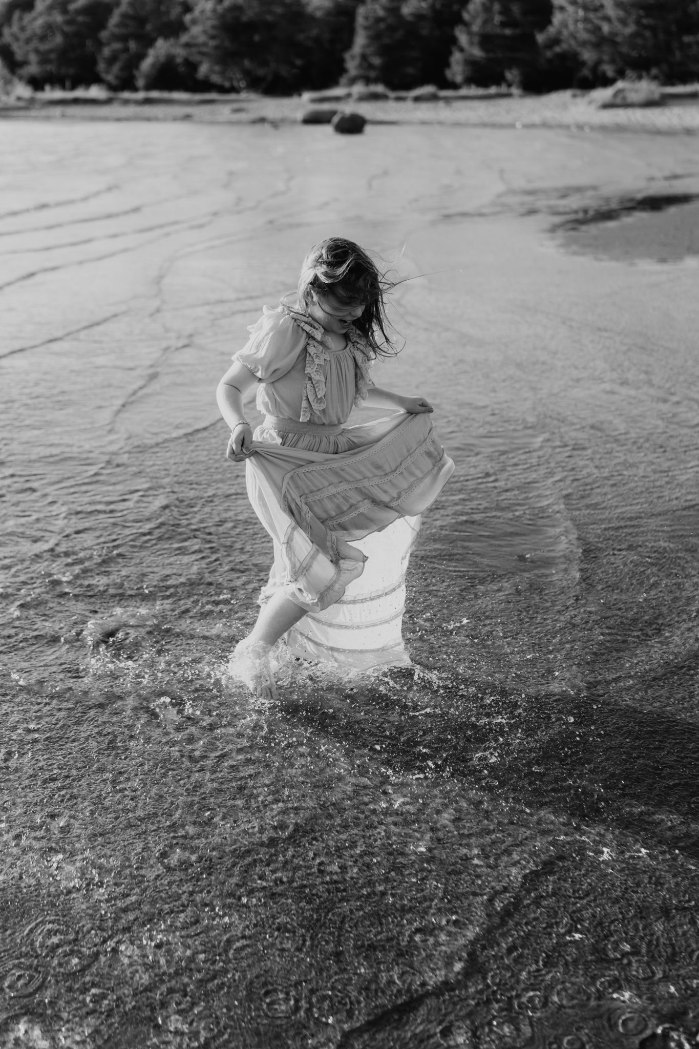 Black and white image of a girl in a flowing dress wading through shallow beach water at sunset.