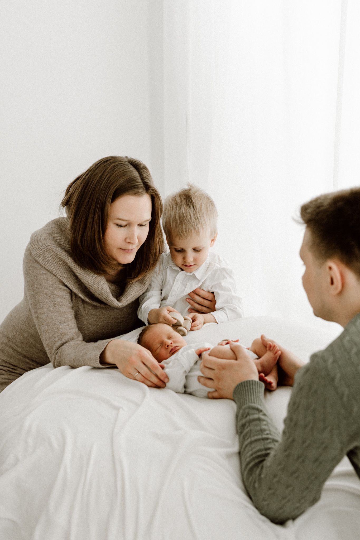 Family with mother, father, and toddler son gathered on white bed admiring their newborn baby.