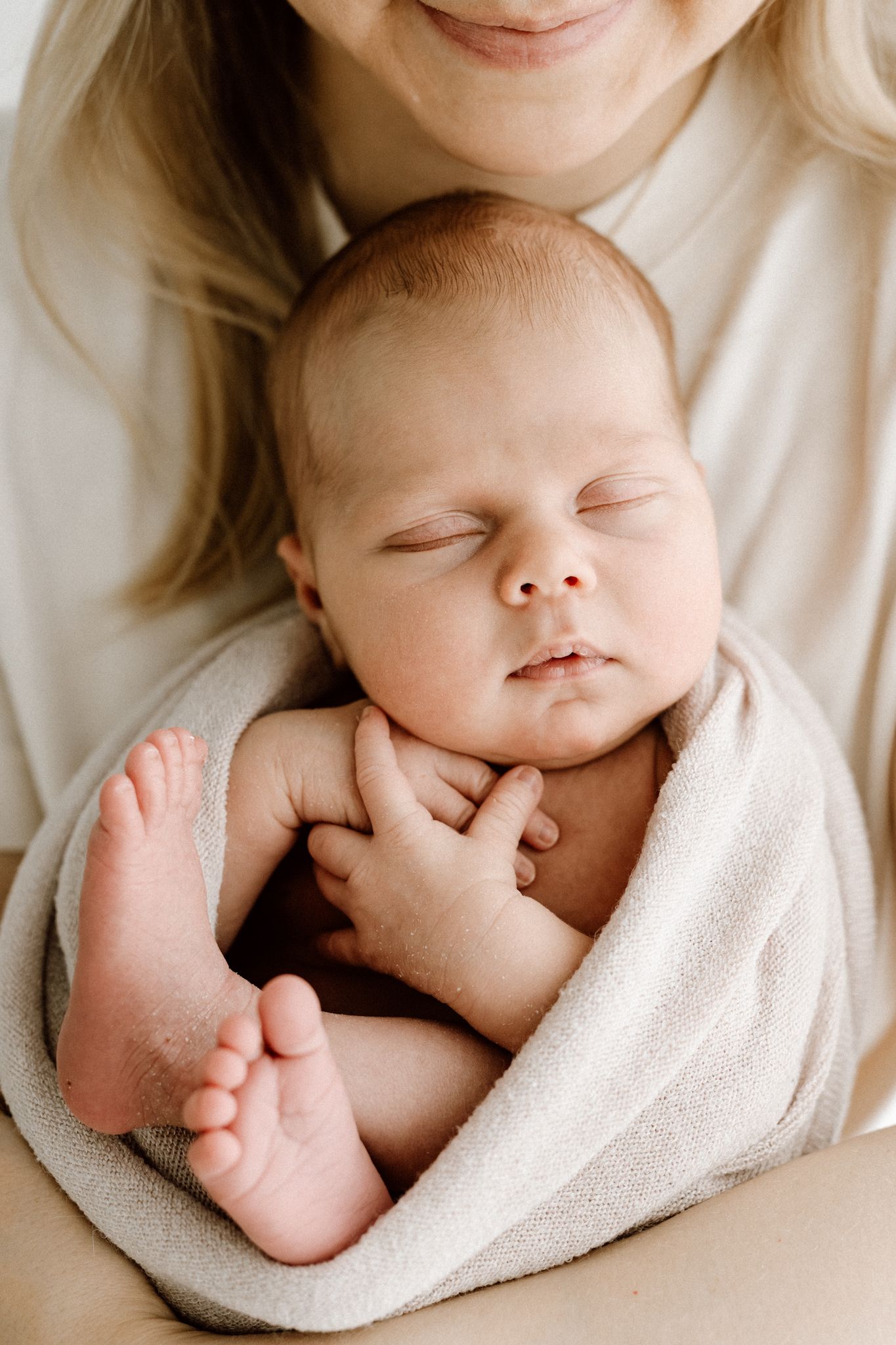 Smiling mother cradling sleeping newborn baby wrapped in soft beige blanket.