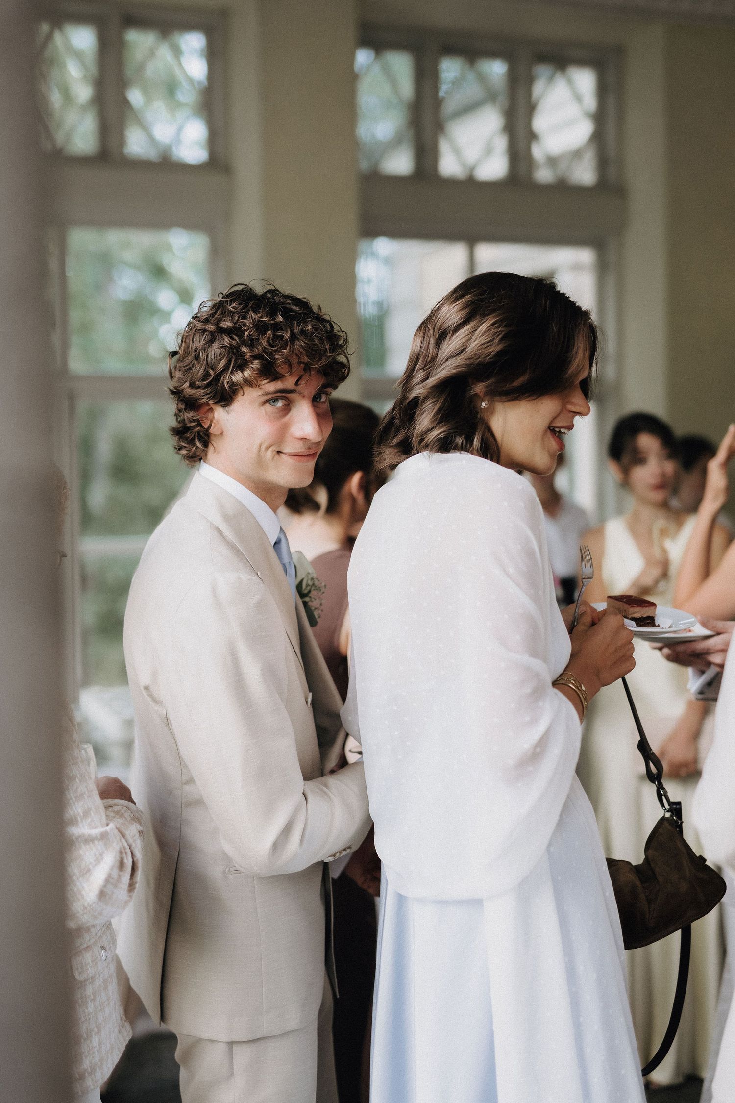 Young groom in beige suit smiles at camera during wedding reception while bride in white stands nearby.