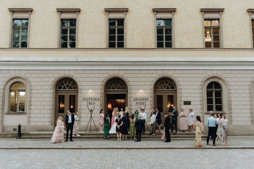 Elegantly dressed wedding guests mingling outside a classical stone building facade.