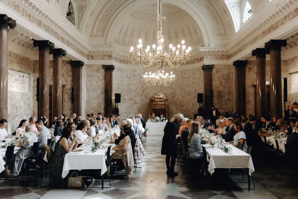 Elegant wedding reception dinner in grand neoclassical ballroom with marble columns and chandeliers.