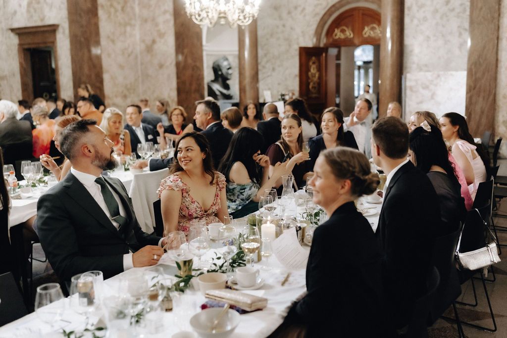 Wedding guests laughing and celebrating at elegantly decorated reception tables in a grand marble ballroom.
