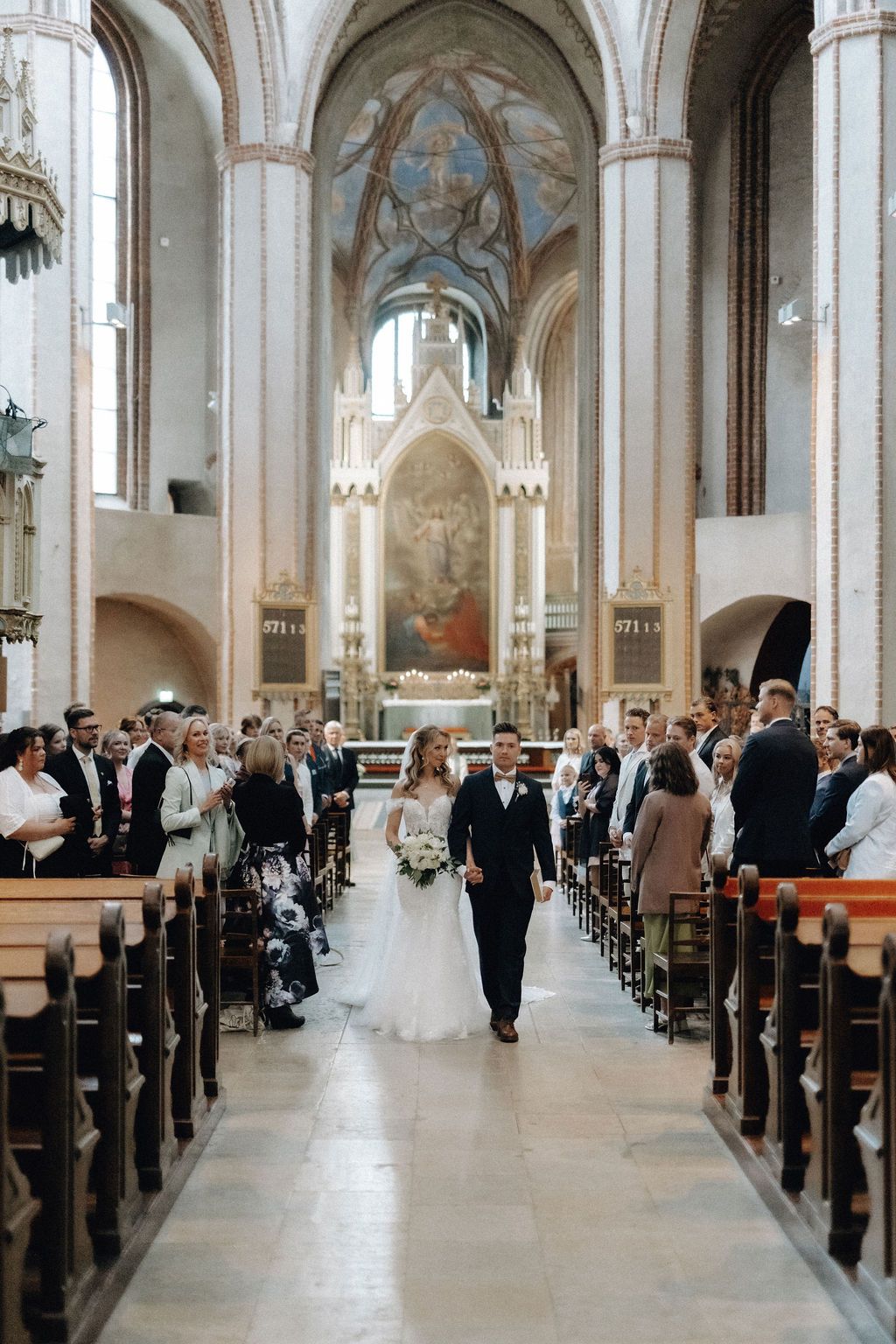 Newlyweds walk hand in hand down church aisle after ceremony in stunning Gothic cathedral with frescoes.