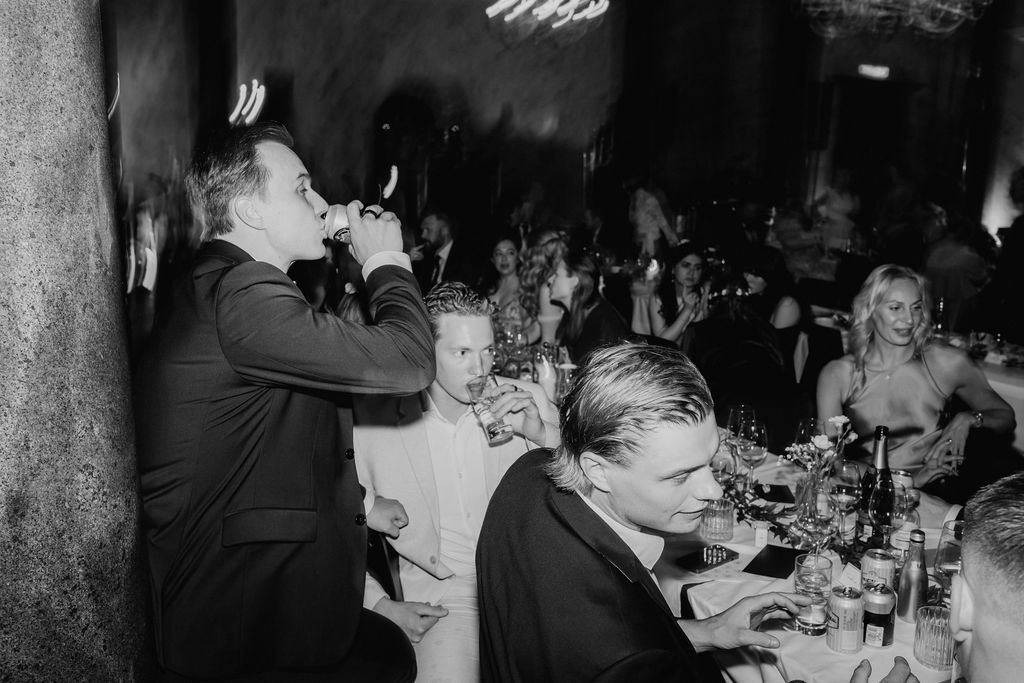 Man in tuxedo drinks while guests celebrate at elegant candlelit wedding reception dinner.