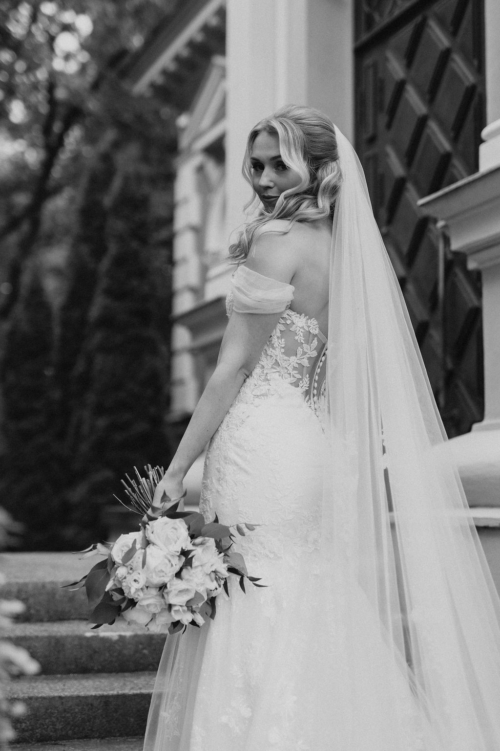 Black and white portrait of bride in lace off-shoulder gown holding bouquet on steps of classical building.