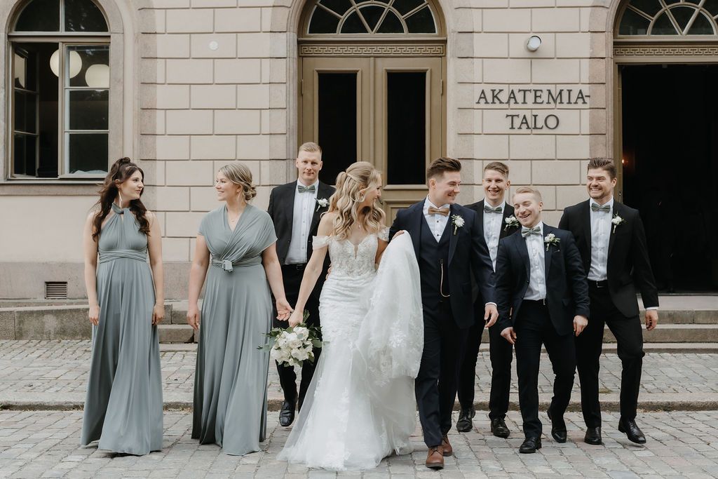 Bride, groom, bridesmaids in sage gowns, and groomsmen pose outside Akatemia-Talo venue.