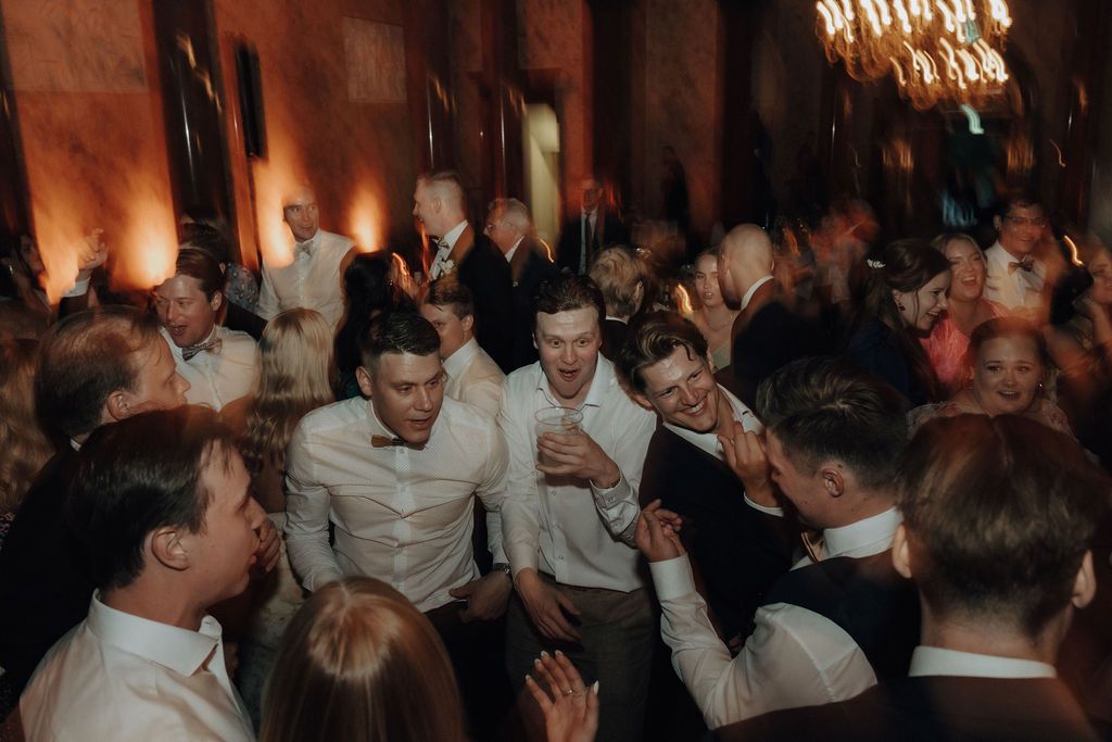 Groomsmen and wedding guests dancing energetically together in an elegant warmly lit ballroom.