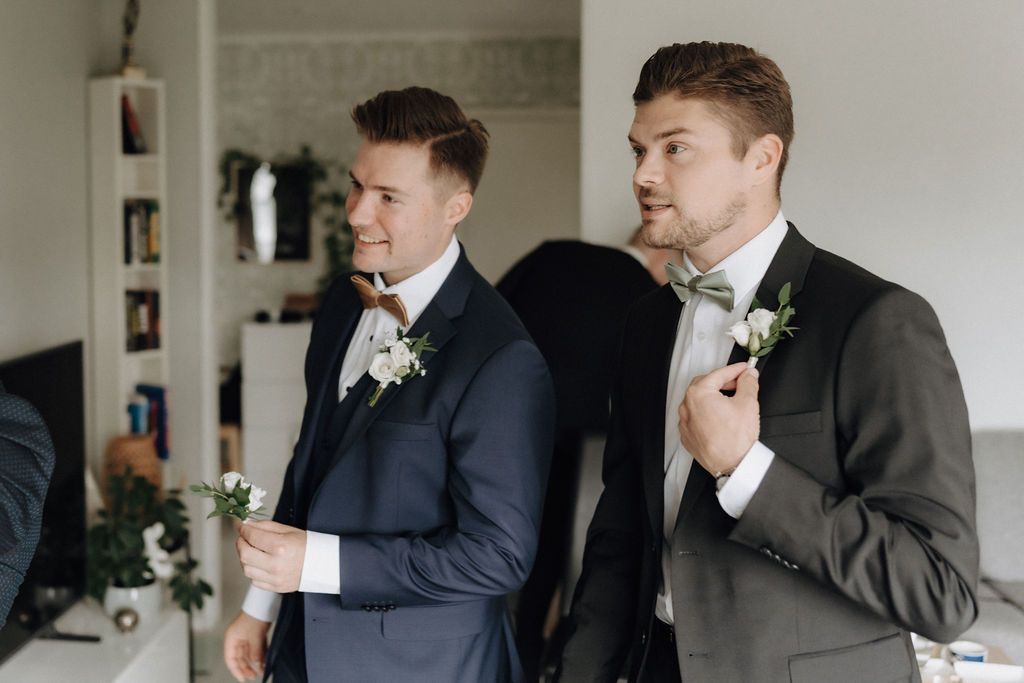Two groomsmen in suits with boutonnieres prepare for a wedding ceremony indoors.