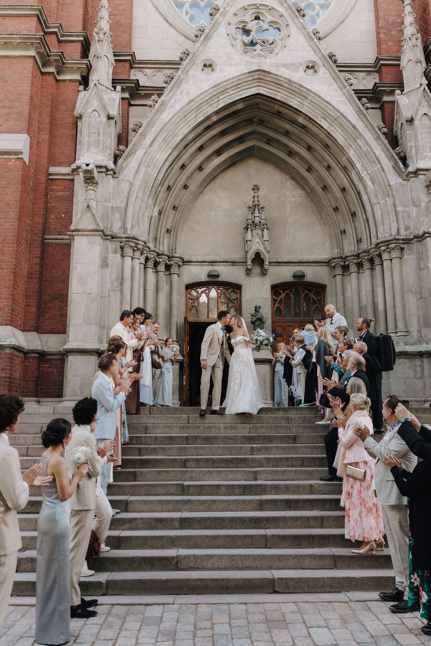 Newlyweds share a kiss on church steps surrounded by applauding guests at a Gothic cathedral.
