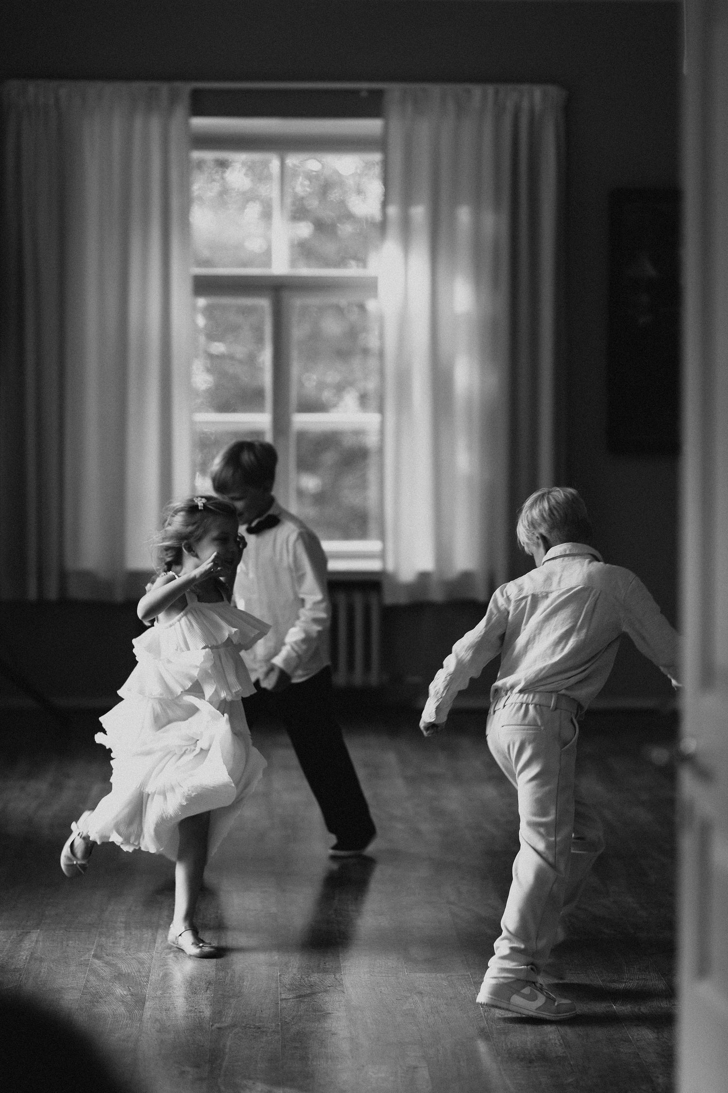 Black and white photo of children dancing joyfully in a sunlit ballroom at a wedding reception.