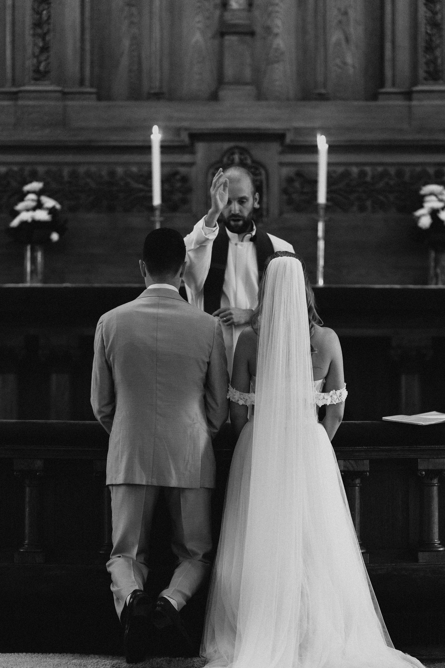 Black and white photo of officiant blessing bride and groom kneeling at ornate wooden church altar with candles.
