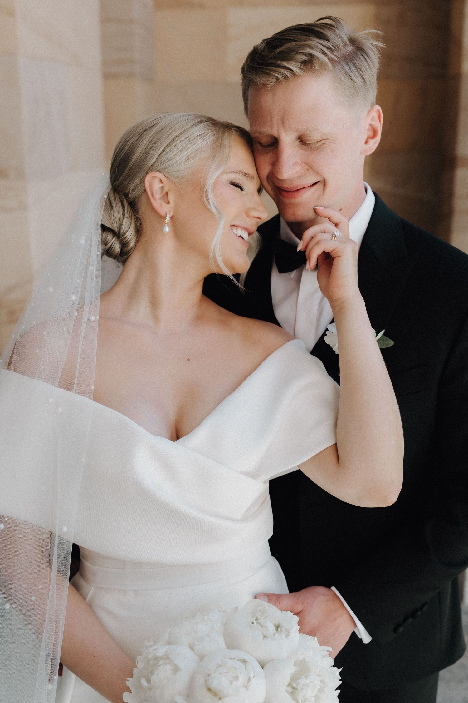 Smiling bride in off-shoulder gown leans into groom in tuxedo while holding white peony bouquet.