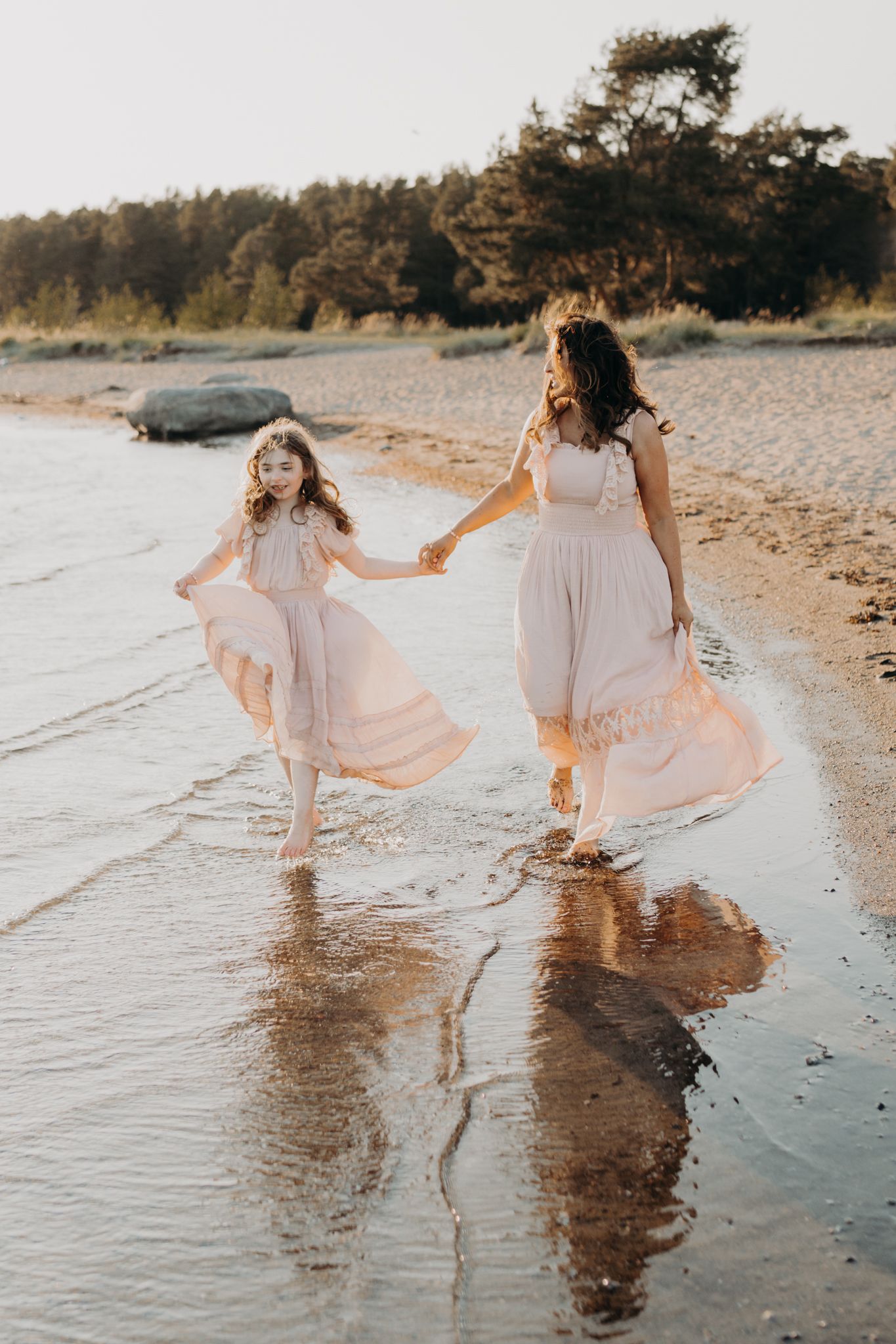A mother and daughter in matching blush pink dresses run hand in hand along a sunlit sandy shoreline.