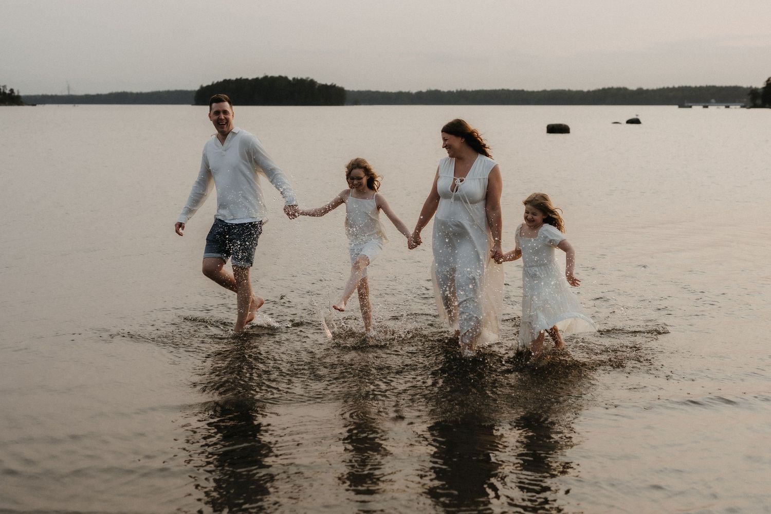 A happy family of four splashes through shallow lake water holding hands at sunset.