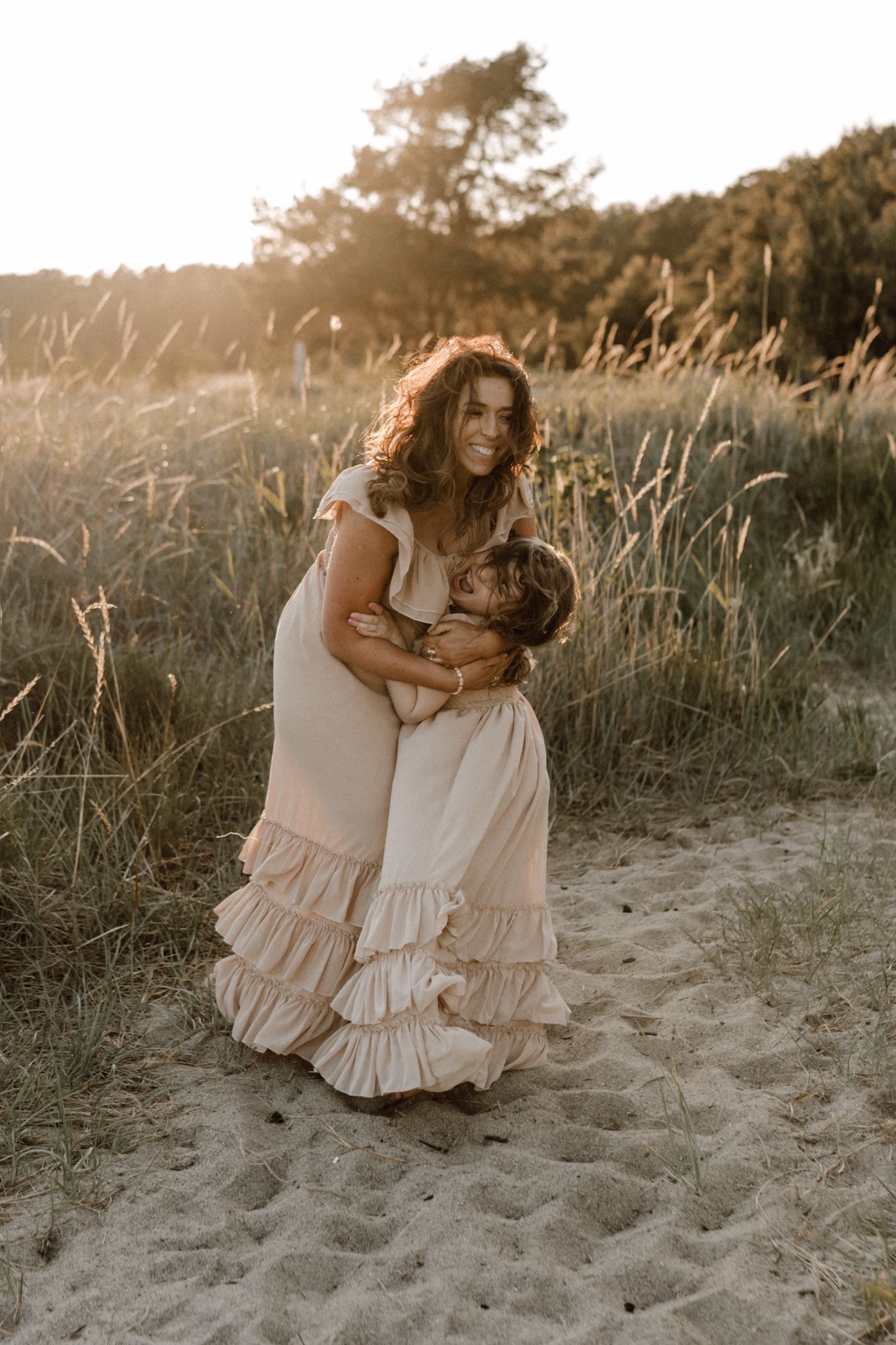 A smiling mother in a ruffled blush dress lovingly embraces her child in a golden sunlit field.