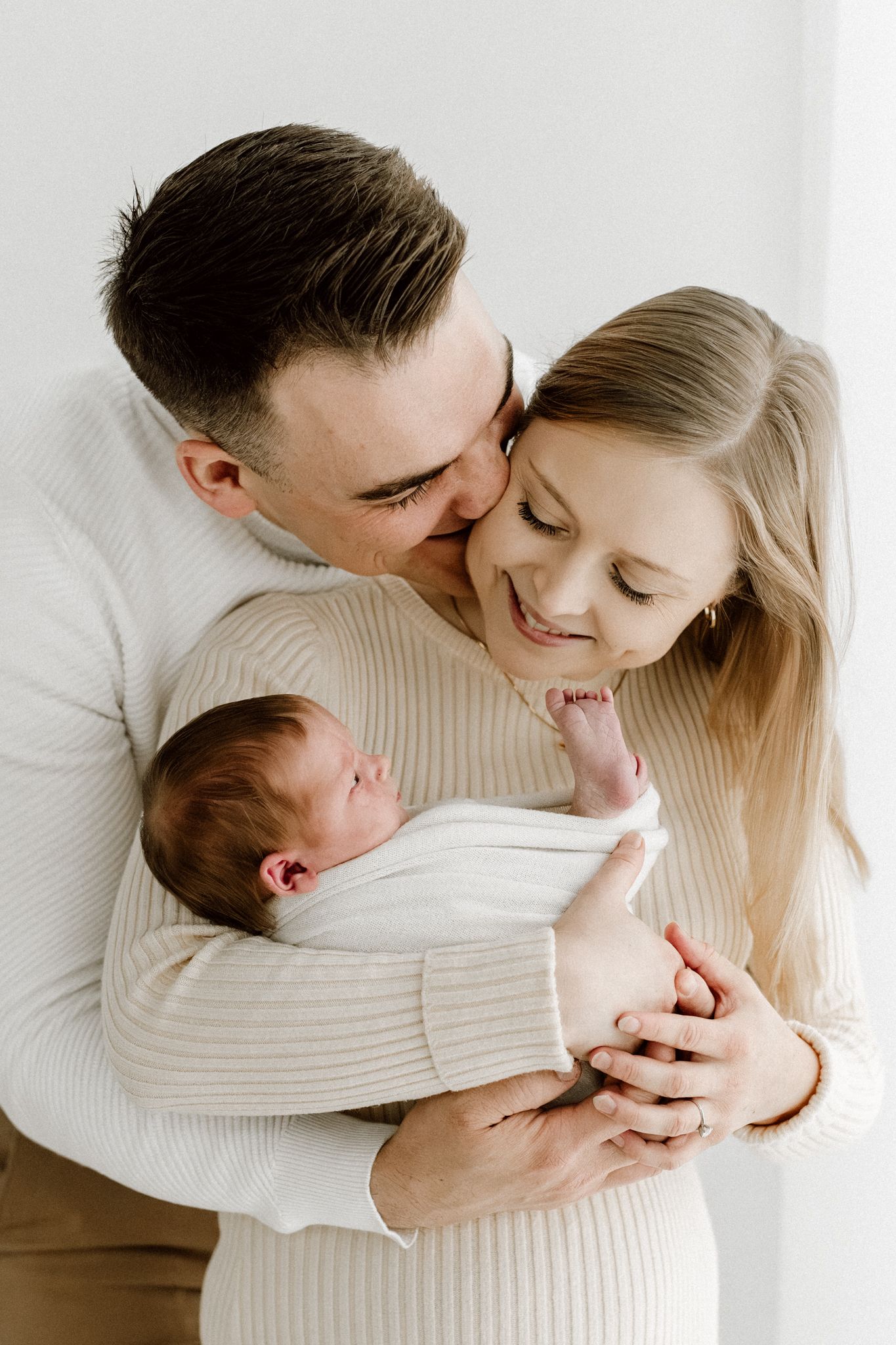 Happy parents in white sweaters tenderly hold their sleeping newborn baby during a professional family photo session.