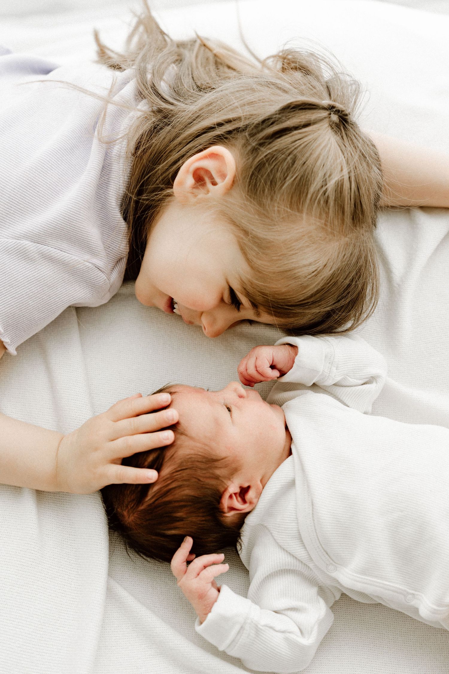 Young girl lying on white bed face-to-face with laughing newborn baby sibling.