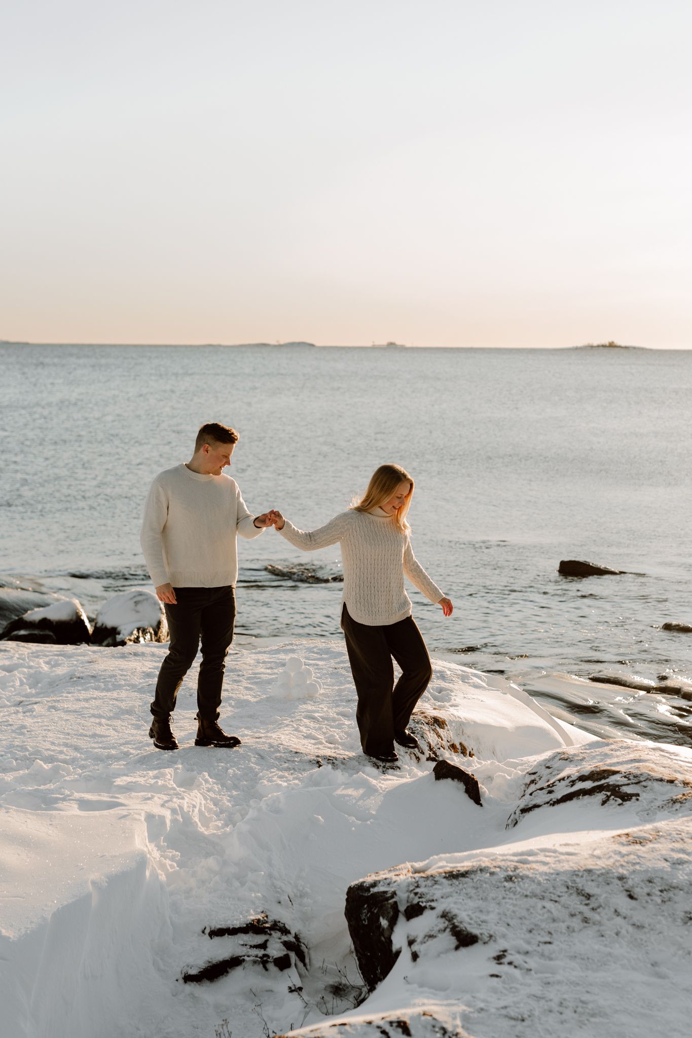 Couple in cream sweaters hold hands while walking on snow-covered rocky shoreline at sunset.