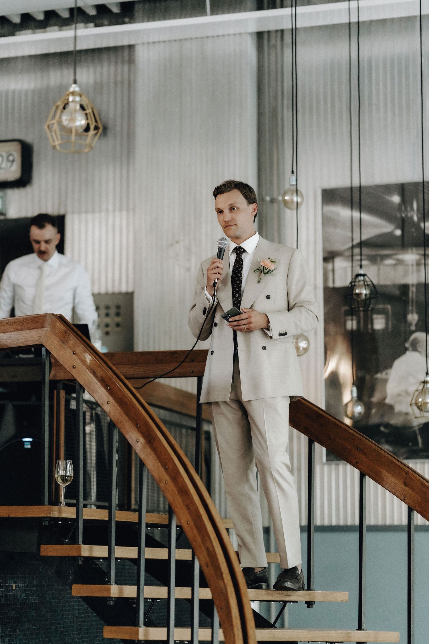 Groom in beige suit giving speech on staircase at wedding reception venue.