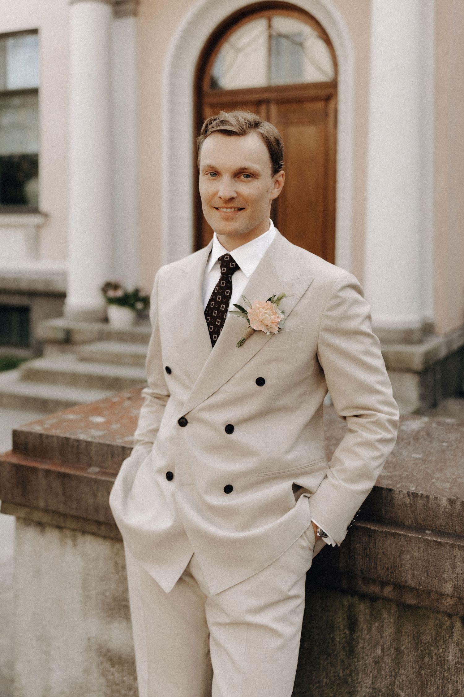 Smiling groom in a beige double-breasted suit with a pink boutonniere and dark floral tie at a classical venue.