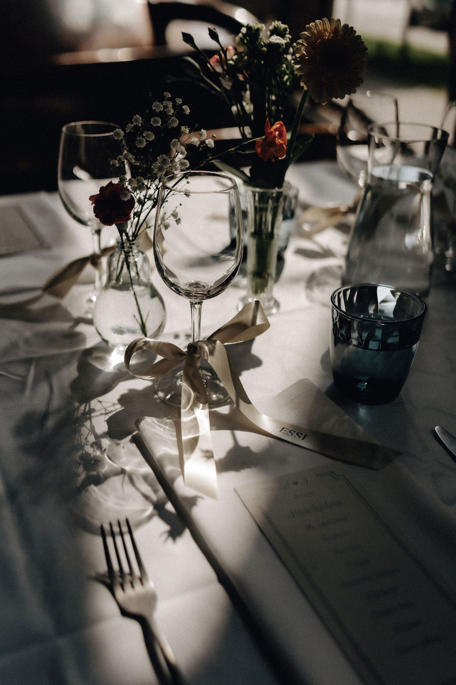 Wedding table detail with wine glasses, ribbon-tied cutlery, menu card, and floral centerpiece in moody light.