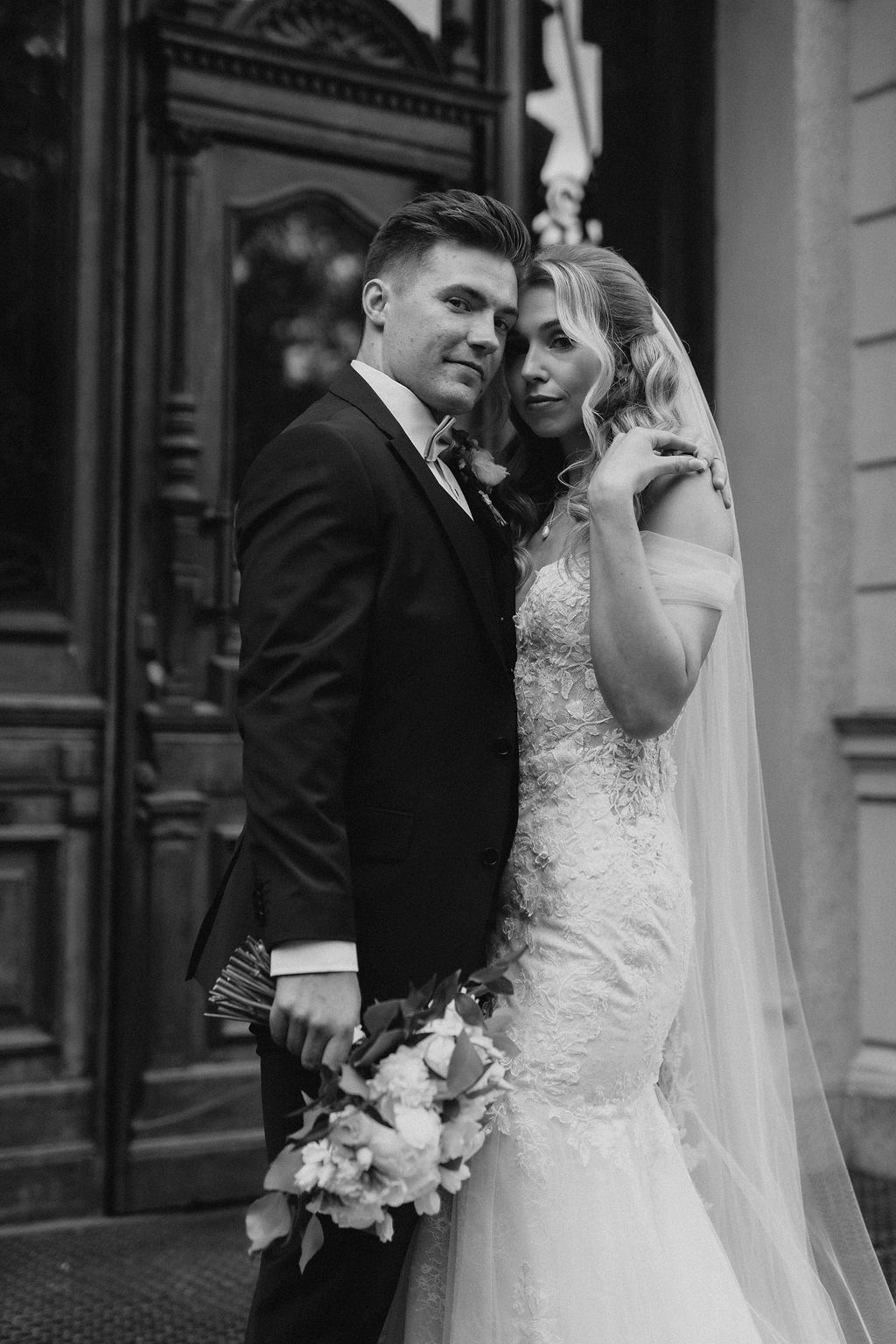 Black and white portrait of bride and groom embracing in front of ornate wooden door of classical building.