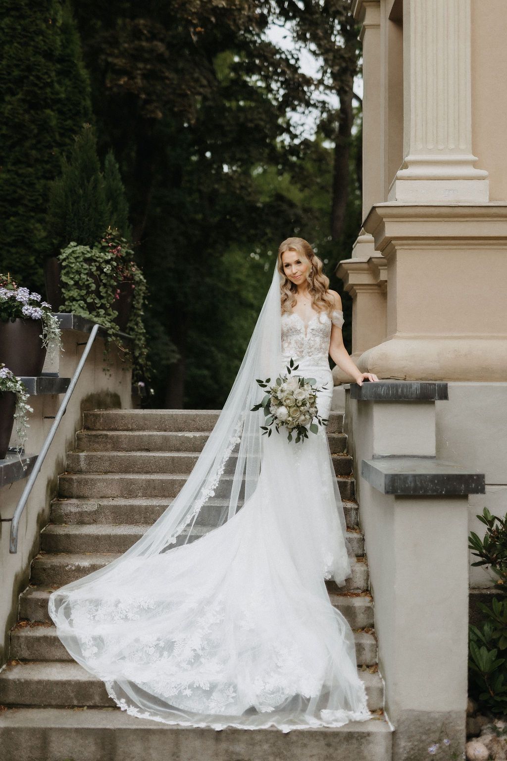 Elegant bride in lace mermaid gown with cathedral veil poses alone on stone steps of classic building.