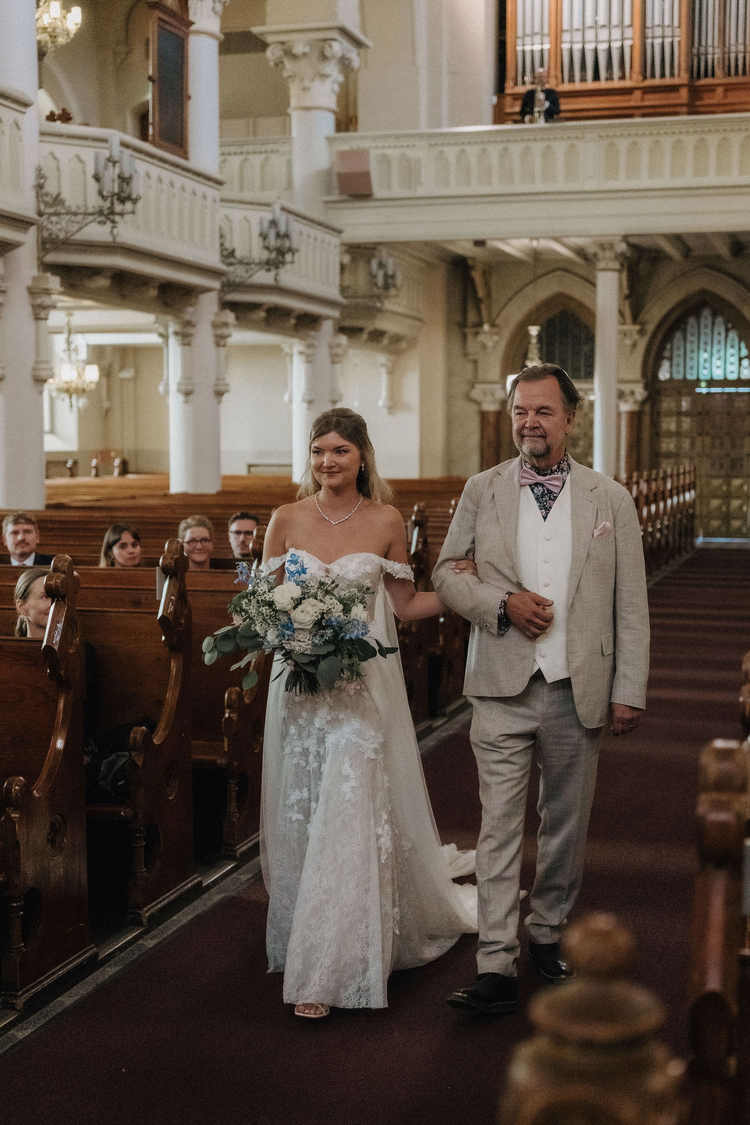 Bride in lace off-shoulder gown walking down the church aisle escorted by her father.