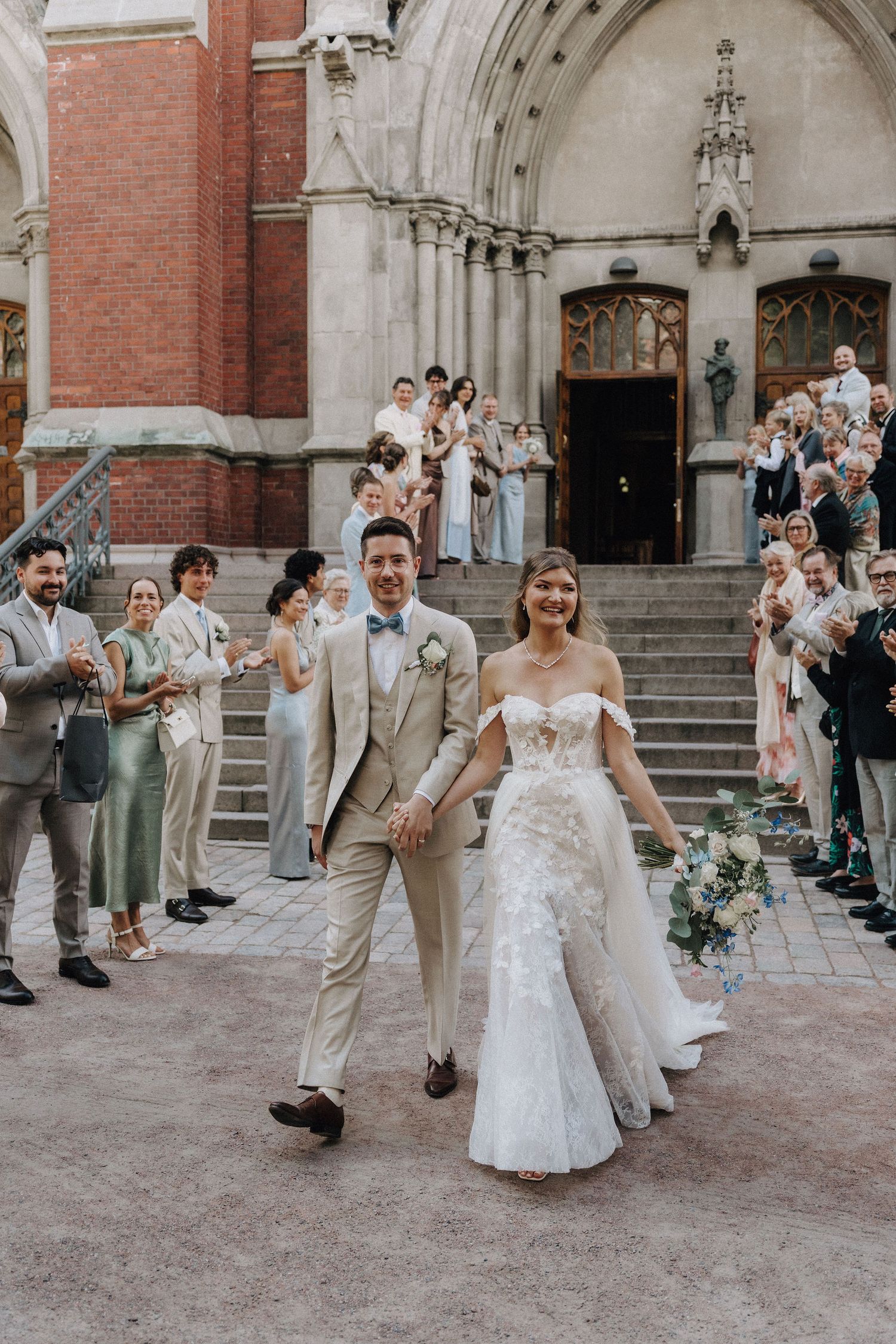 Smiling bride in floral gown and groom in beige suit walk hand in hand outside a Gothic church.