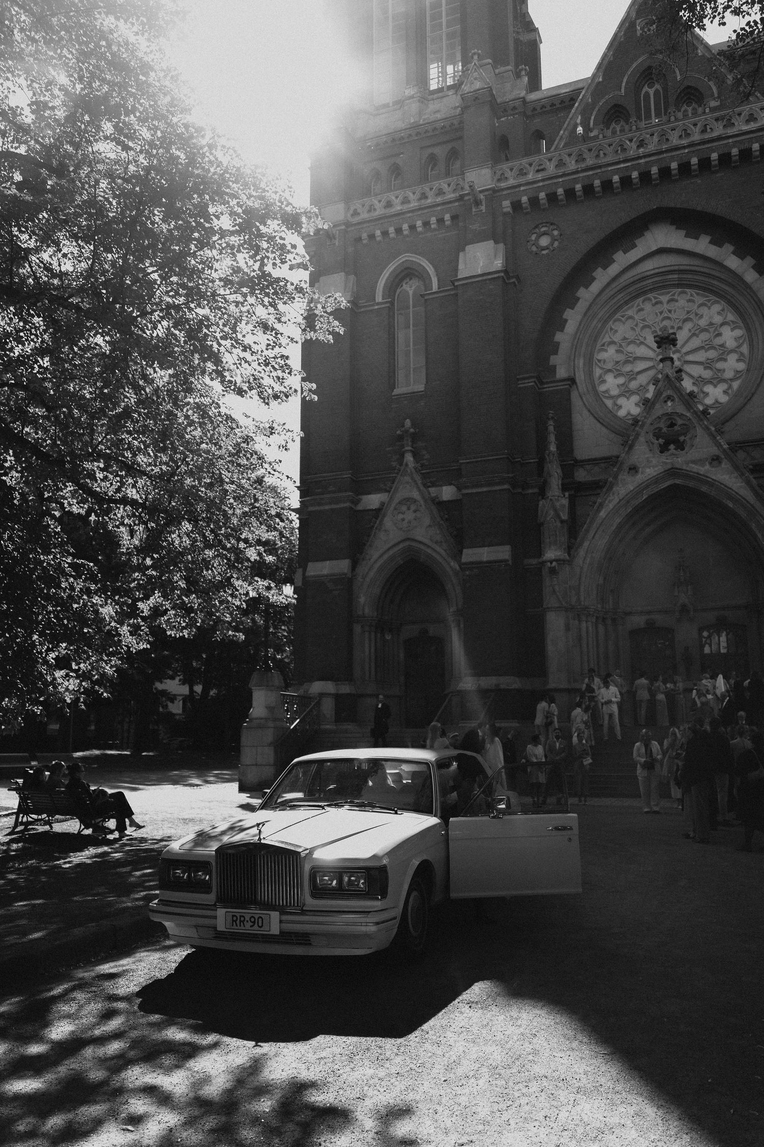 Black and white photo of a white Rolls-Royce parked outside a sunlit Gothic brick church.