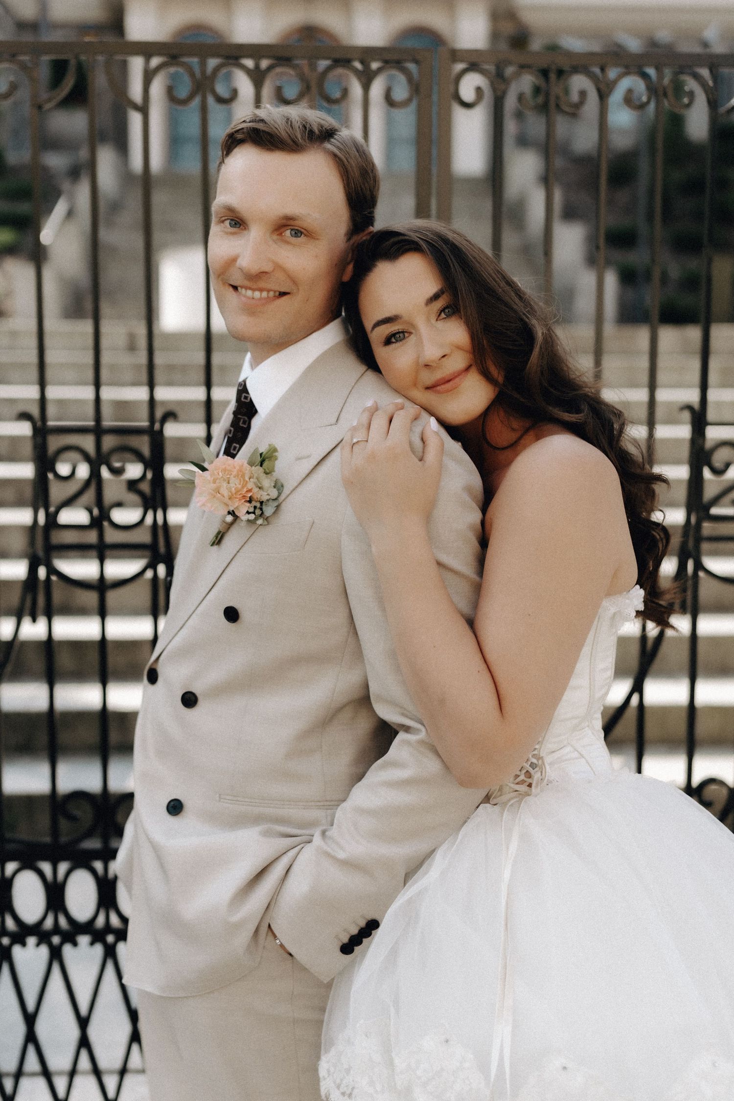 Elegant bride leans on groom's back by ornate iron fence, both smiling softly in classic wedding attire.