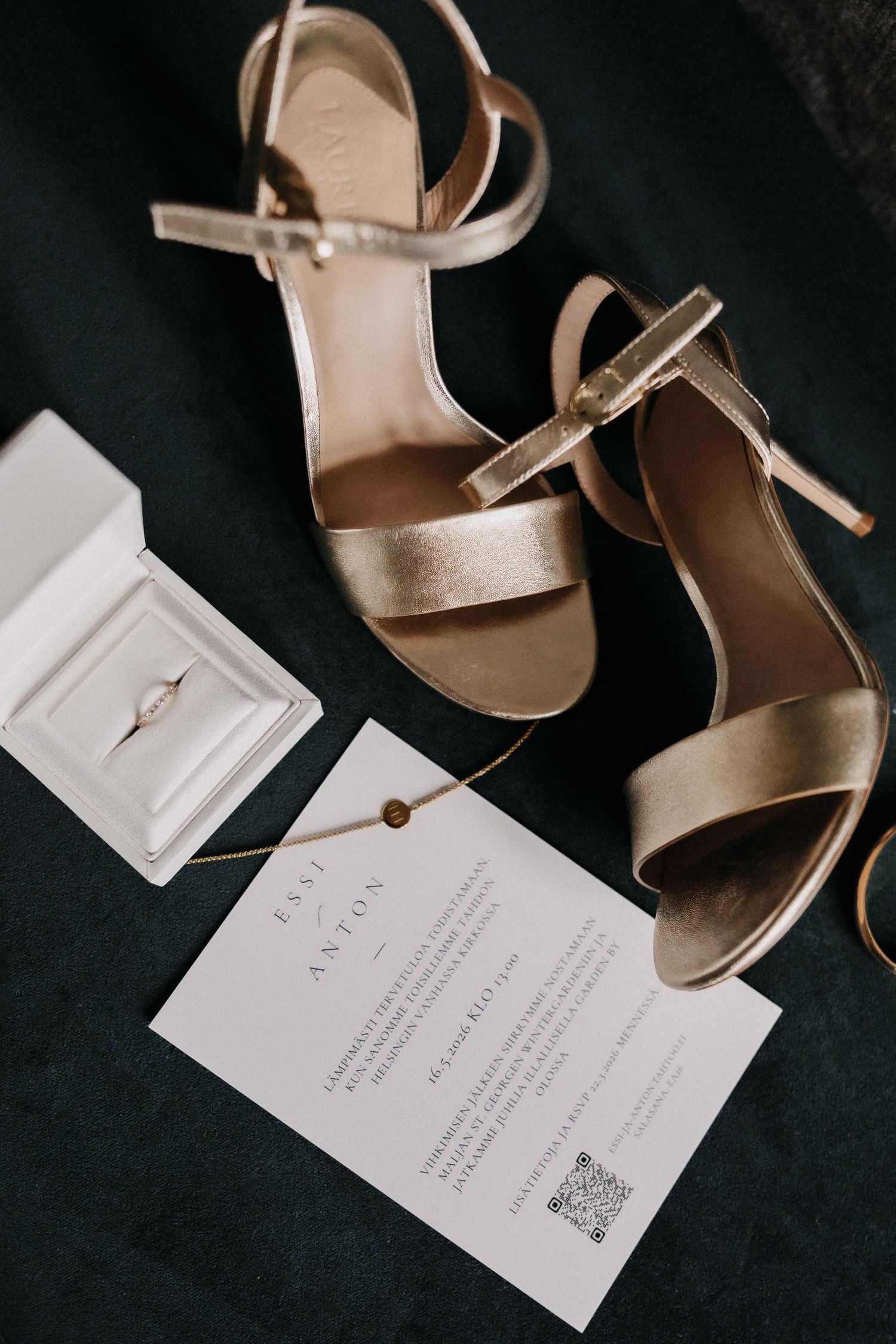 Gold strappy bridal heels, diamond engagement ring, and elegant wedding invitation styled on green velvet.