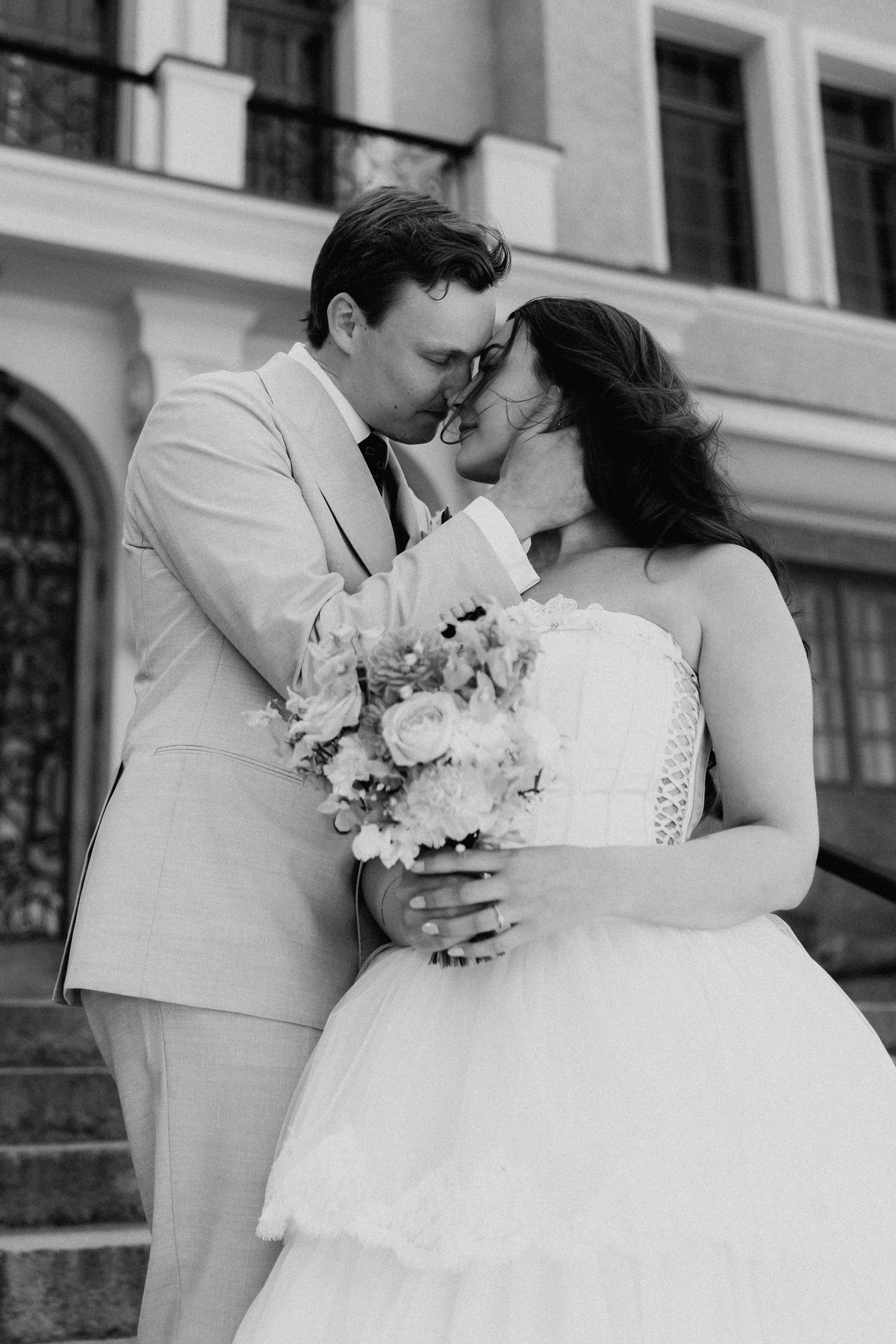 Black and white photo of bride and groom sharing a tender kiss on steps outside an elegant building.