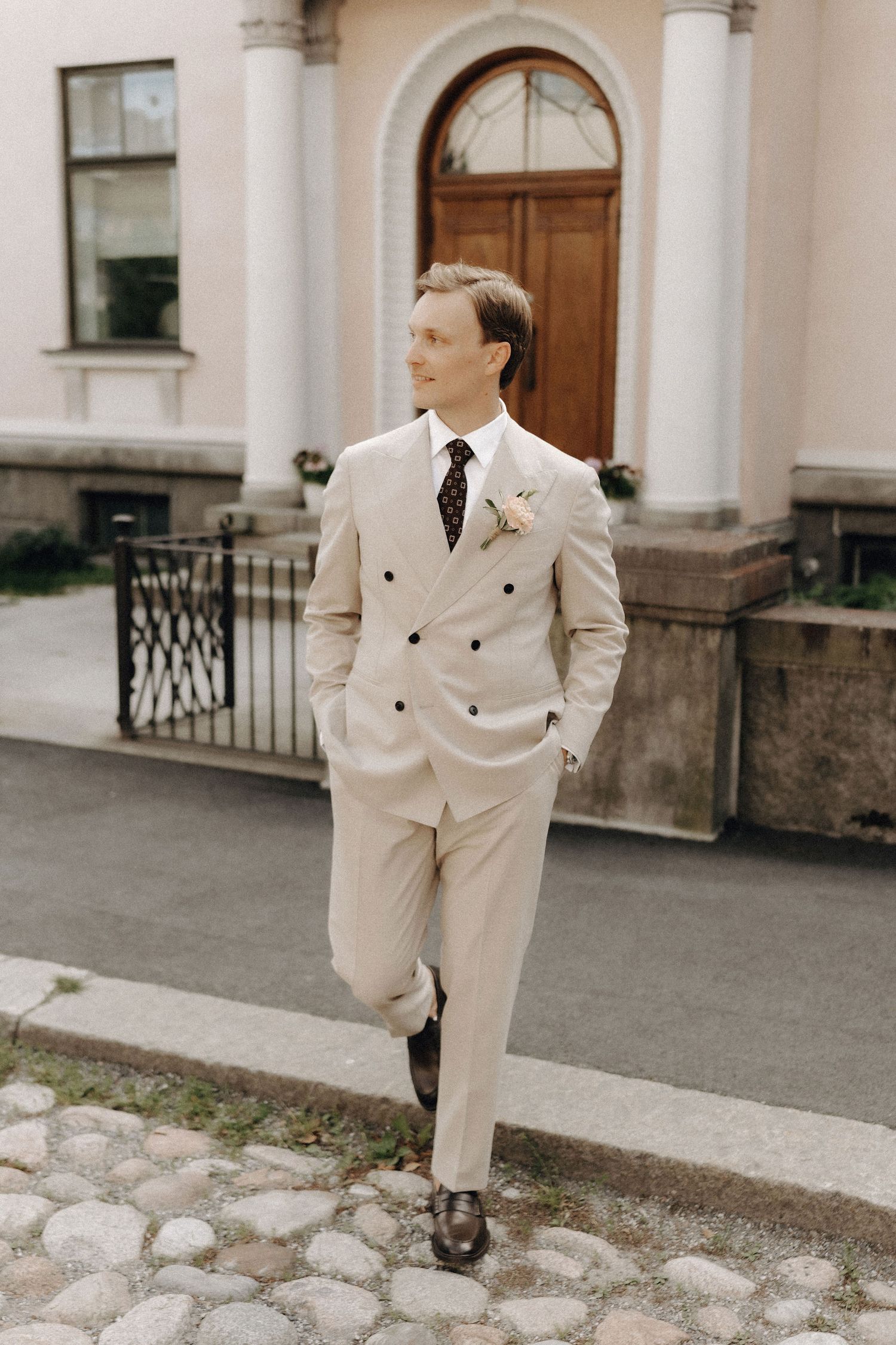Confident groom in a beige double-breasted suit with boutonniere walking on cobblestones outside a classic building.
