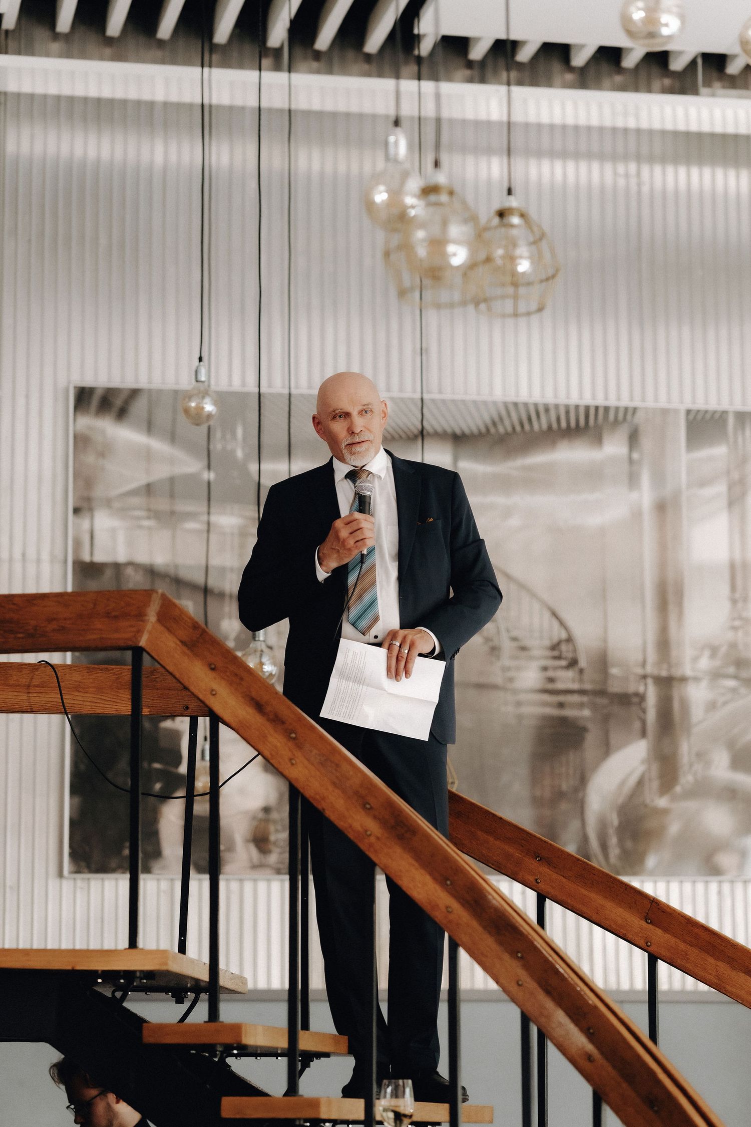 Elderly man in a dark suit delivers a speech on stairs holding a microphone and papers.