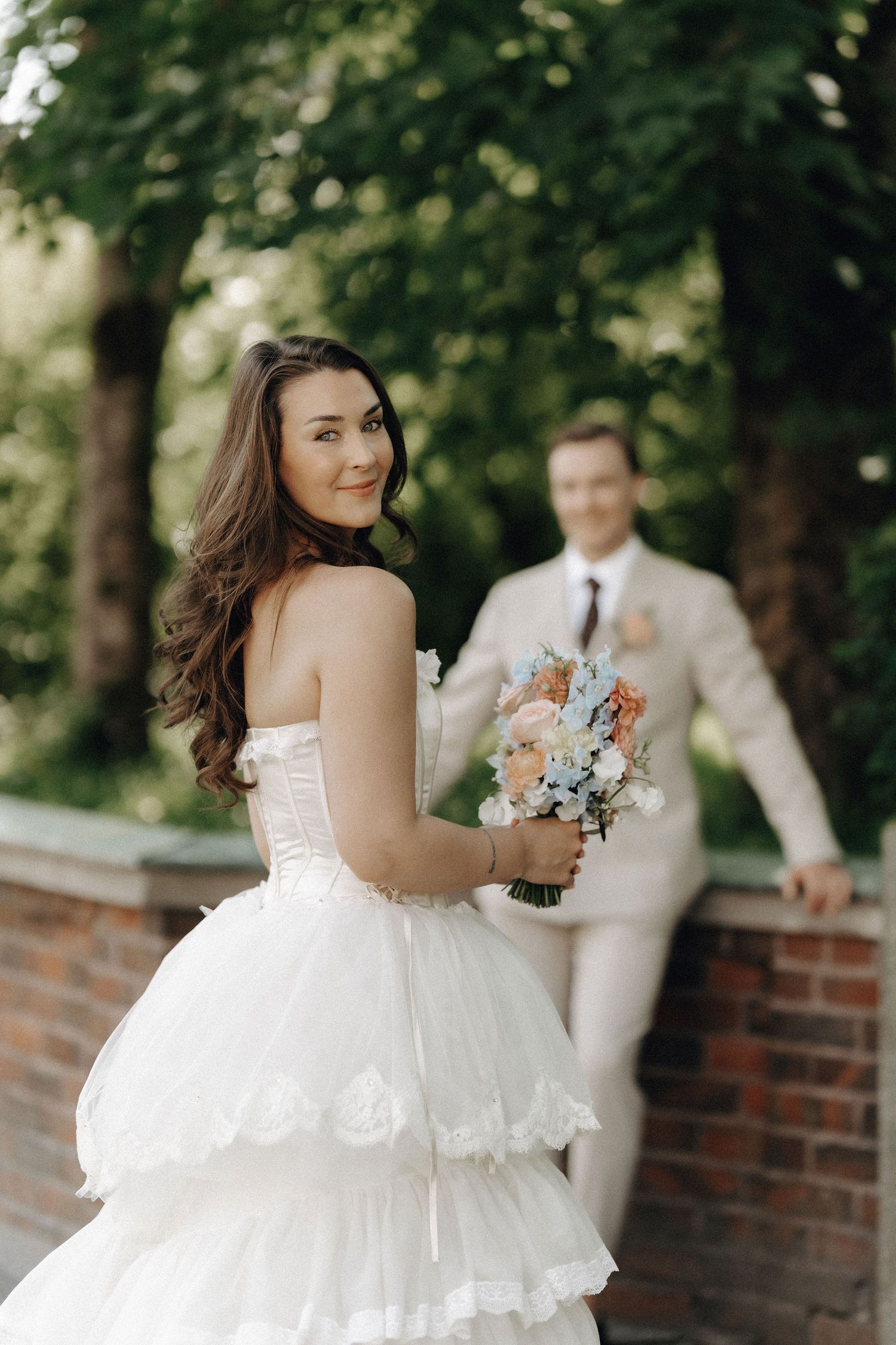 Brunette bride in white ruffled gown smiles over shoulder holding pastel bouquet with groom behind her.