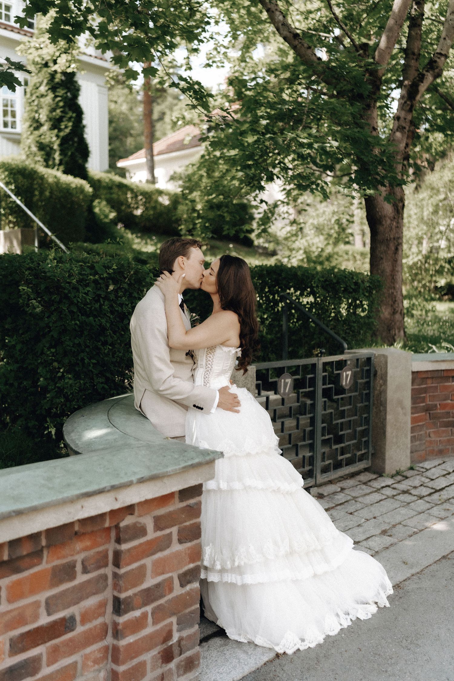 Bride and groom sharing a romantic kiss while seated on a brick wall in lush garden.